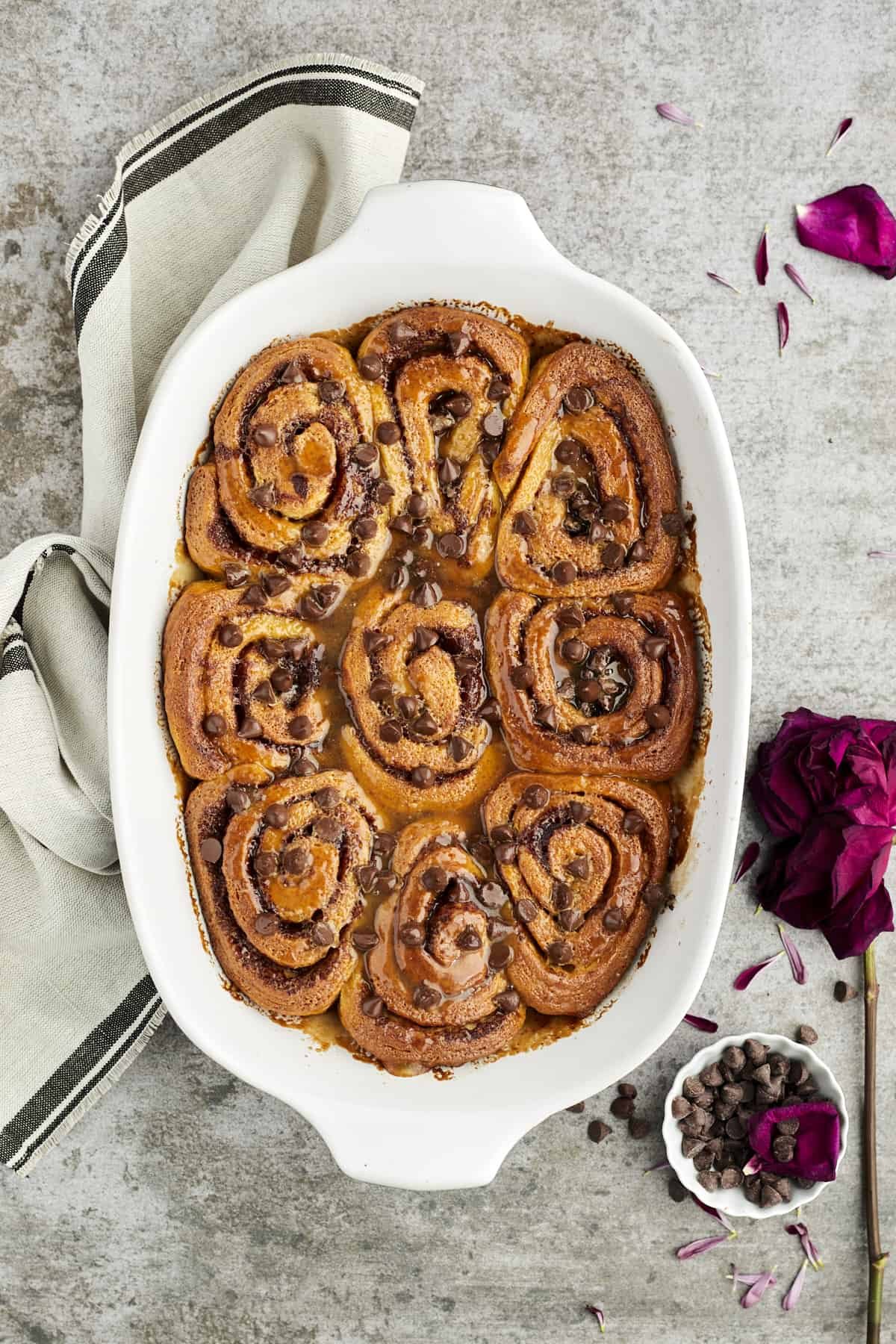 overhead image of simple cinnamon rolls topped with caramel and chocolate chips in a white baking dish with a towel and a small bowl of chocolate chips off to the side