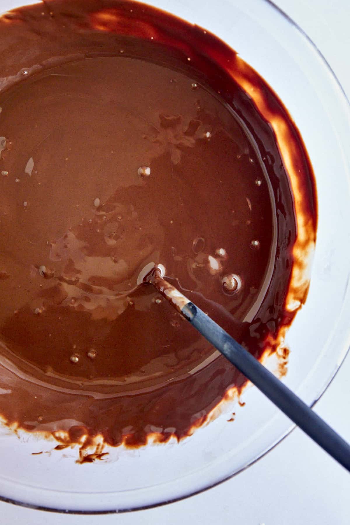 A bowl of melted chocolate and coconut oil with a spoon sticking out.