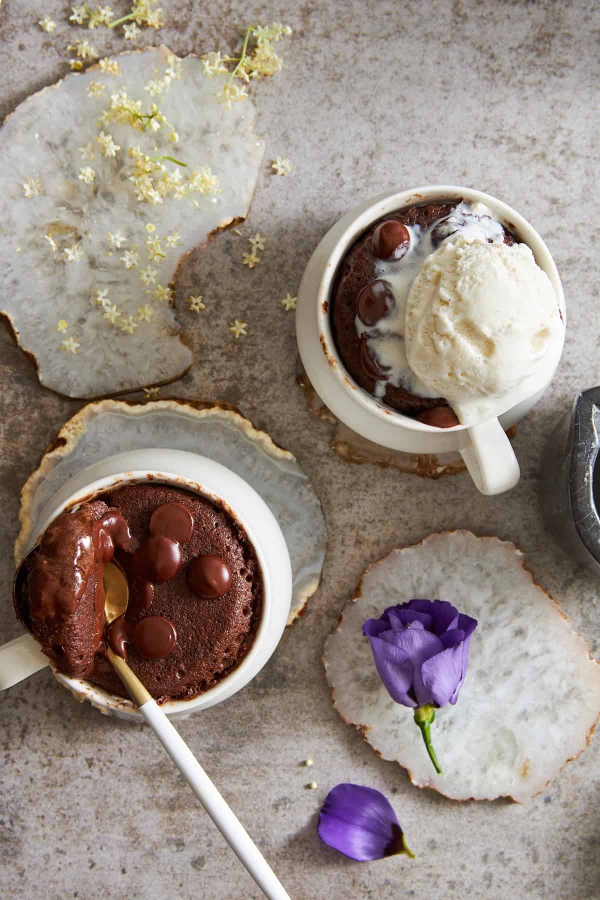 Two chocolate mug cakes, one with a spoon lifting a bite and the other topped with ice cream.