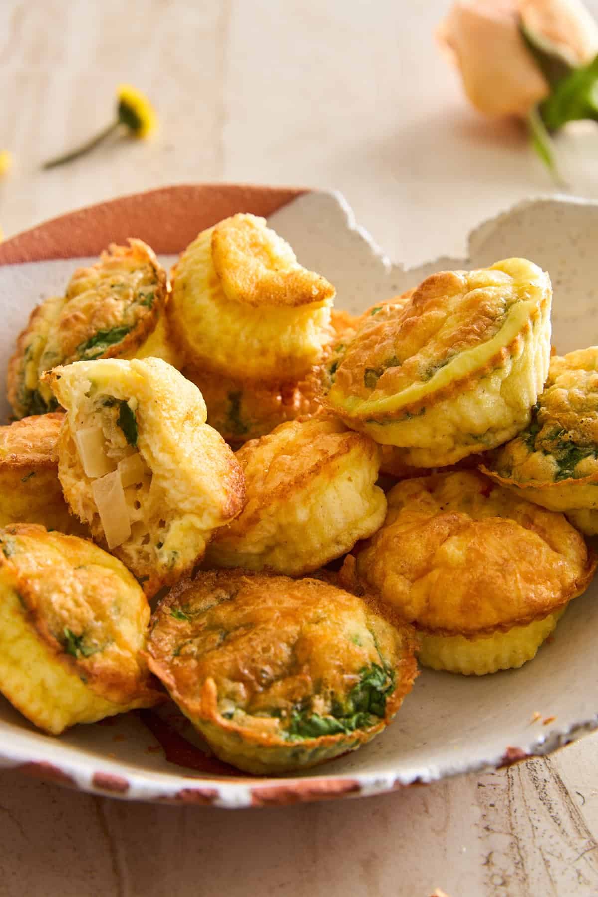 A plate of mini egg muffins with golden-brown tops, filled with spinach and cheese. One muffin is cut open, revealing the fluffy inside and chunky filling. The plate sits on a light surface with flowers in the background.