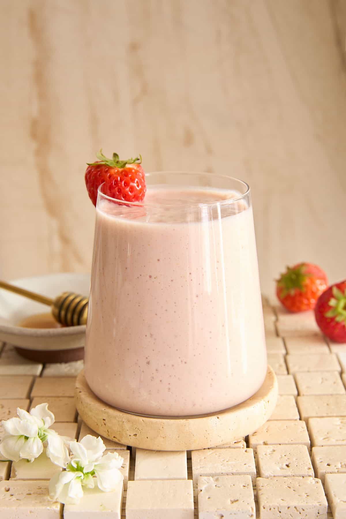A glass of silky strawberry cottage cheese smoothie sits on a round stone coaster, garnished with a whole strawberry. Nearby are two strawberries, a plate with a honey dipper, and white flowers, all on a tiled surface.