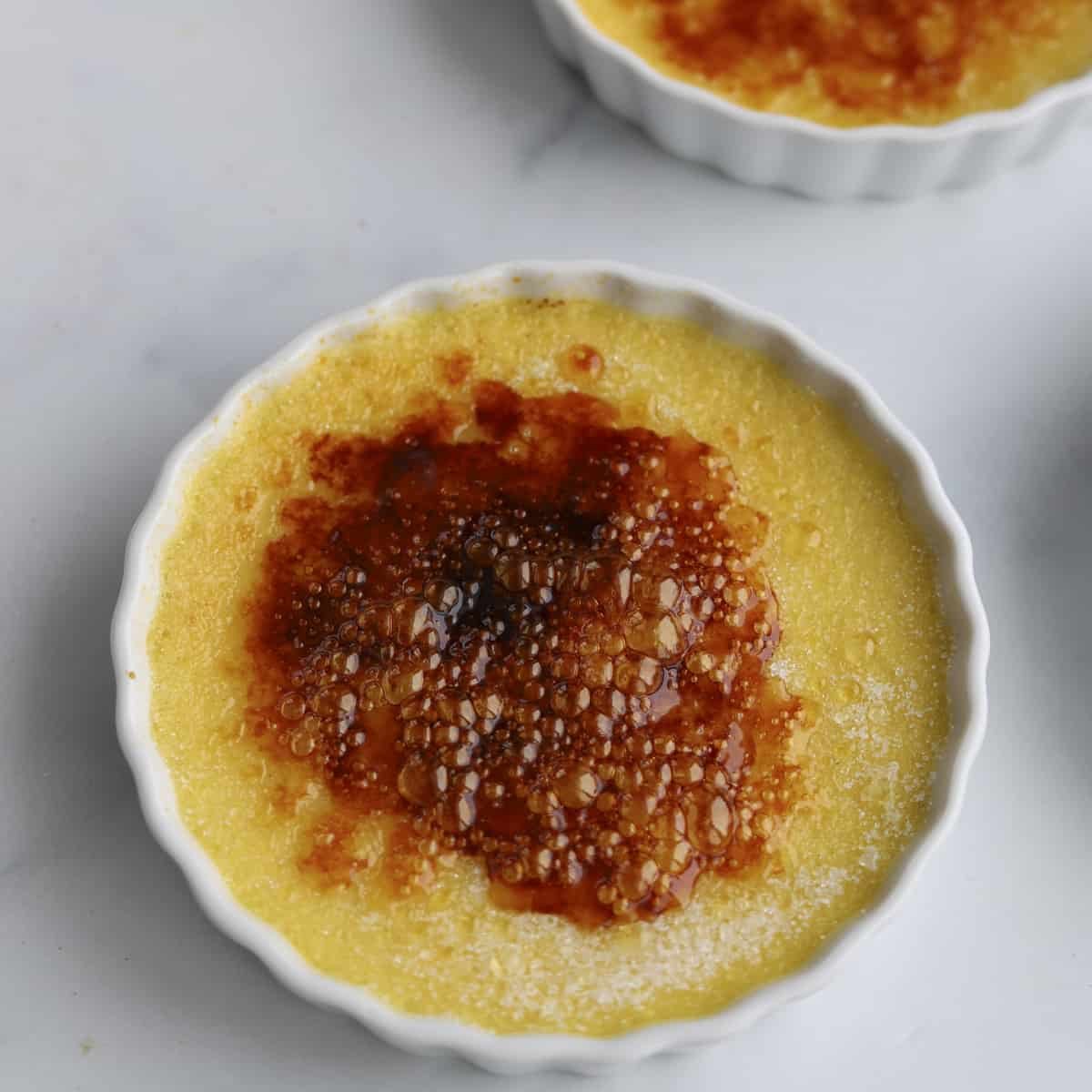 A close-up of a caramelized sugar crust on top of the creamy custard. Another ramekin is partially visible in the background.