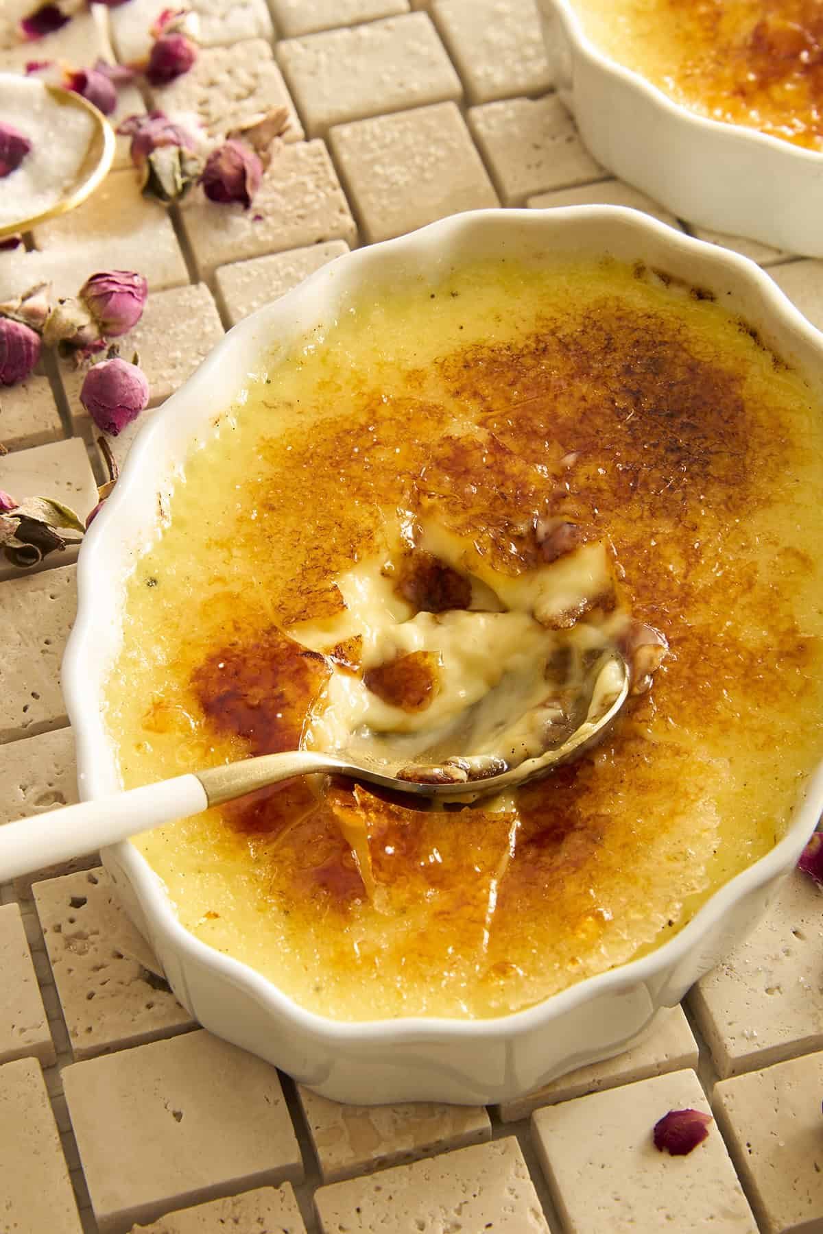 A close-up of a crème brûlée in a white dish, with a spoon breaking through the caramelized sugar crust to reveal the creamy custard underneath. Dried rosebuds are scattered on the tiled surface around the dish.