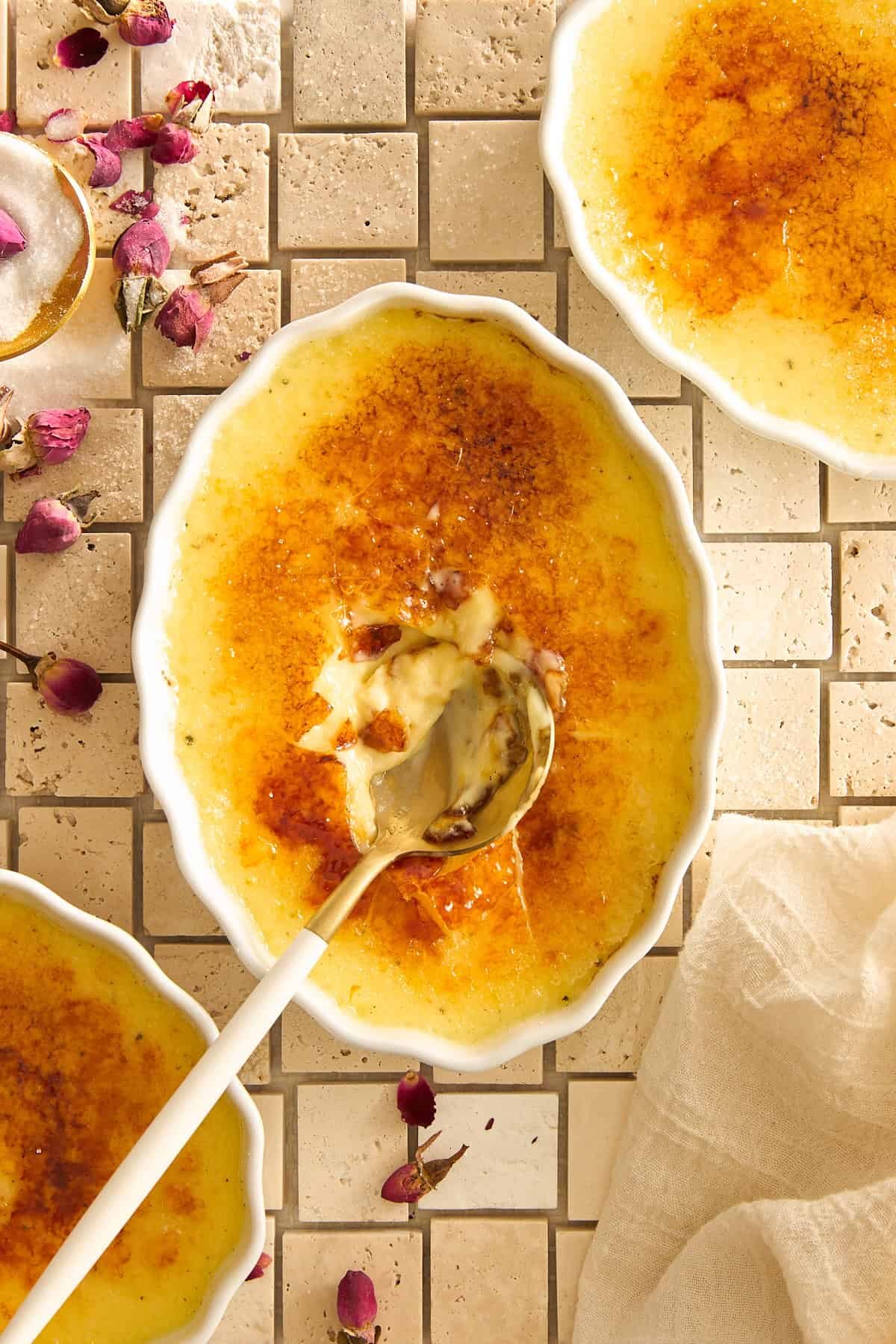 Overhead view of a crème brûlée dish with a caramelized sugar top cracked open by a spoon. Other dishes and scattered dried rosebuds are visible on a cream-colored tile surface. A beige napkin is to the side.