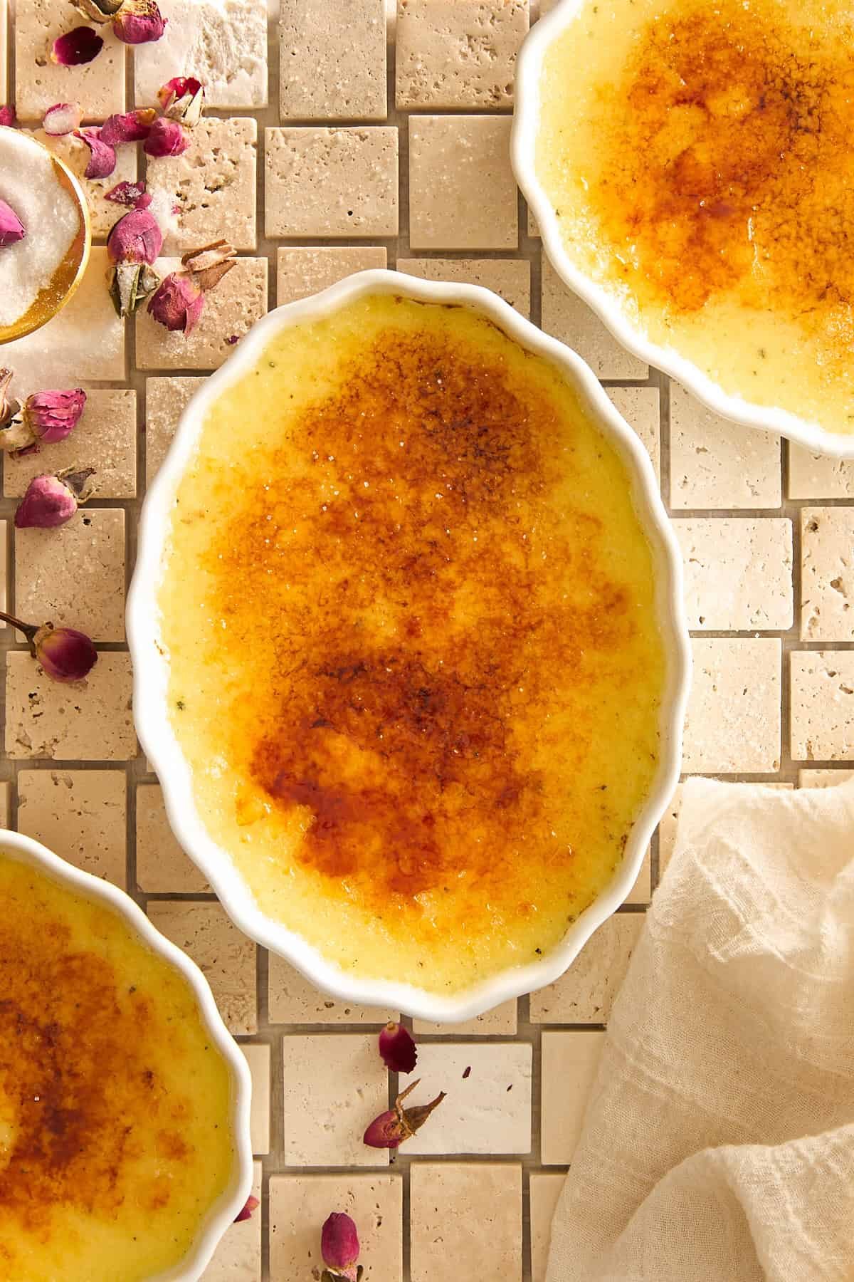 An oval dish of crème brûlée with a caramelized sugar crust sits on a tiled surface, surrounded by scattered dried rosebuds and two partial dishes of crème brûlée. A beige cloth is partially visible in the corner.