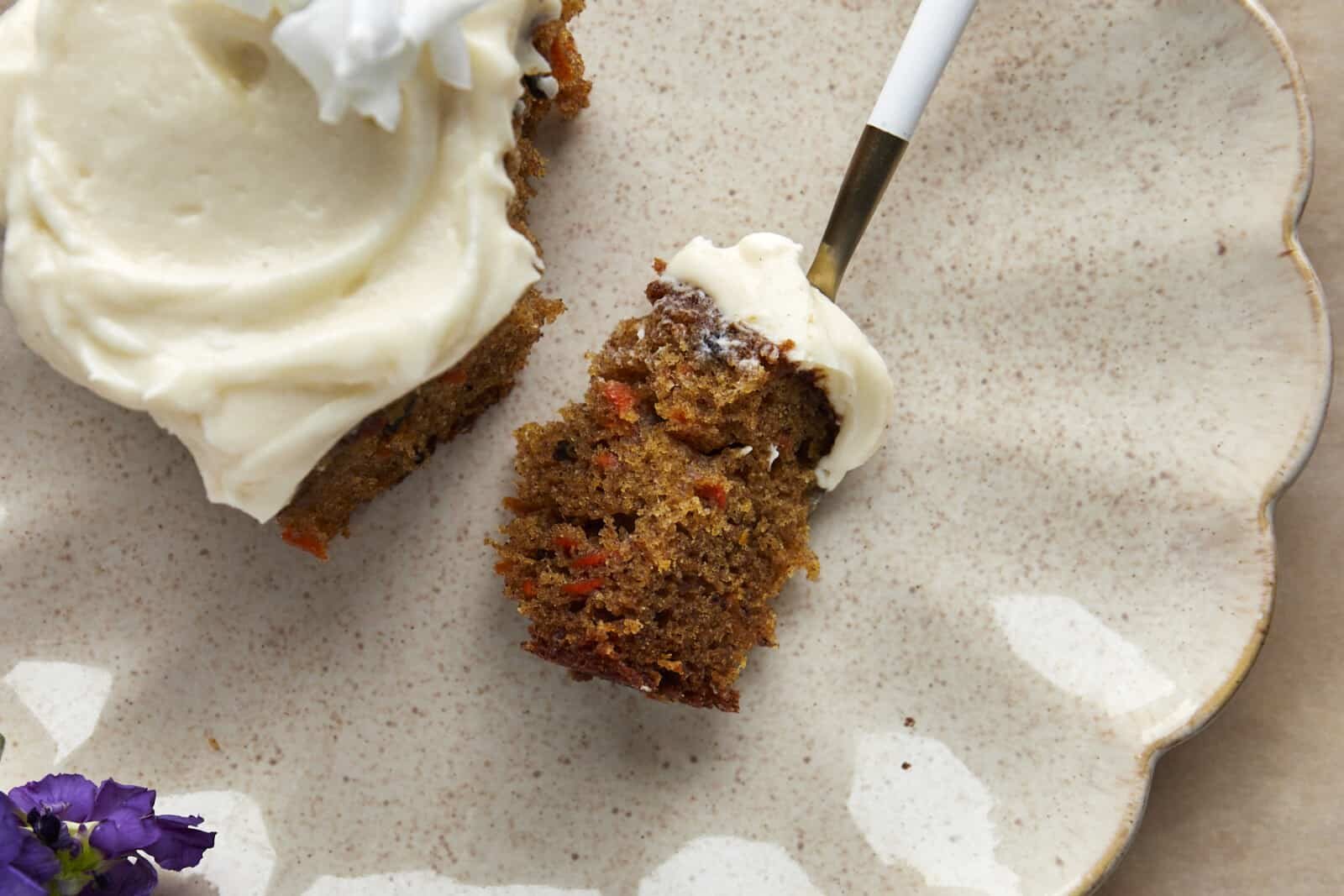 A forkful of carrot cake topped with vanilla bean cream cheese frosting on a plate with the rest of the piece of carrot cake.