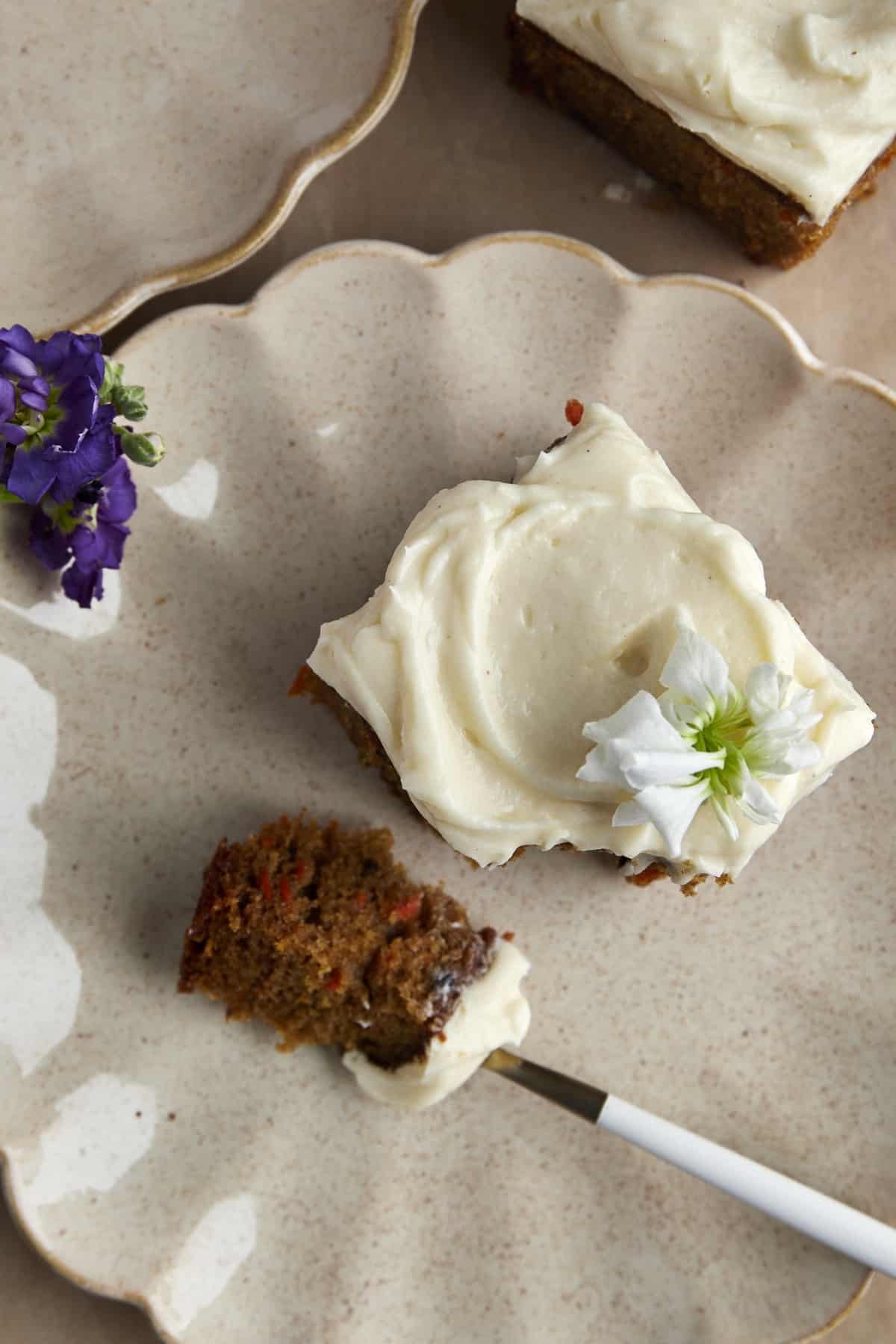 A square of carrot cake topped with cream cheese frosting on a plate with a fork holding a bite.