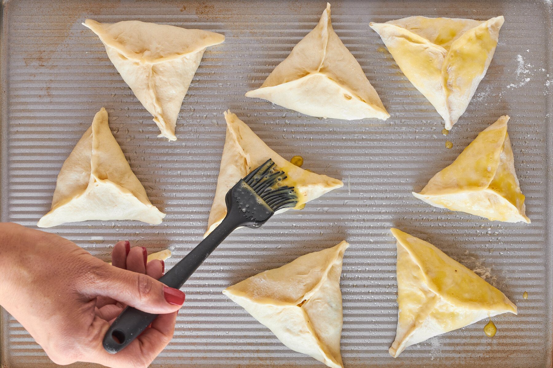 Fatayer on a baking sheet being brushed with egg wash.