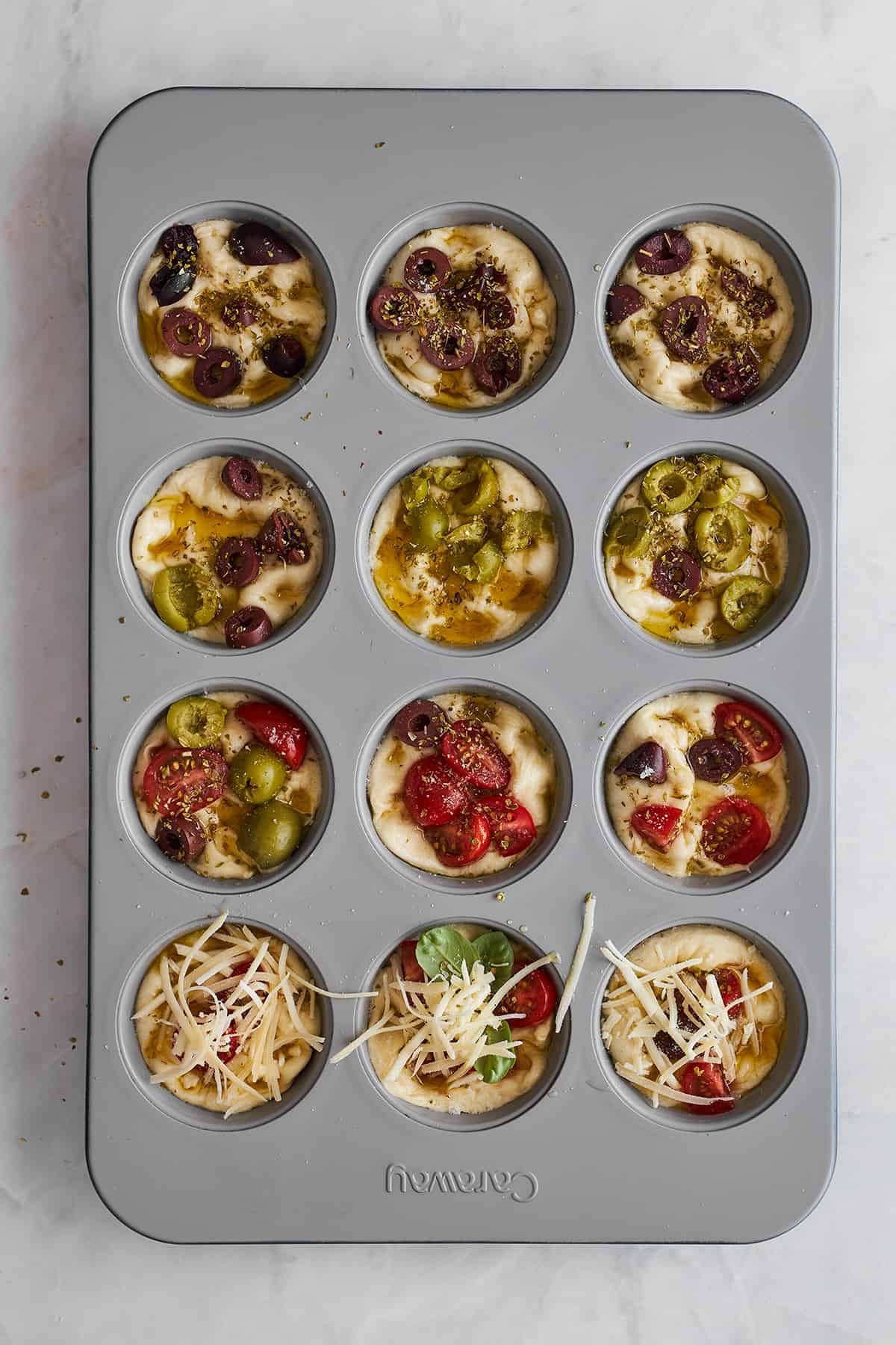 Raw bread focaccia muffins with olives, tomatoes, and cheese in a muffin tin.