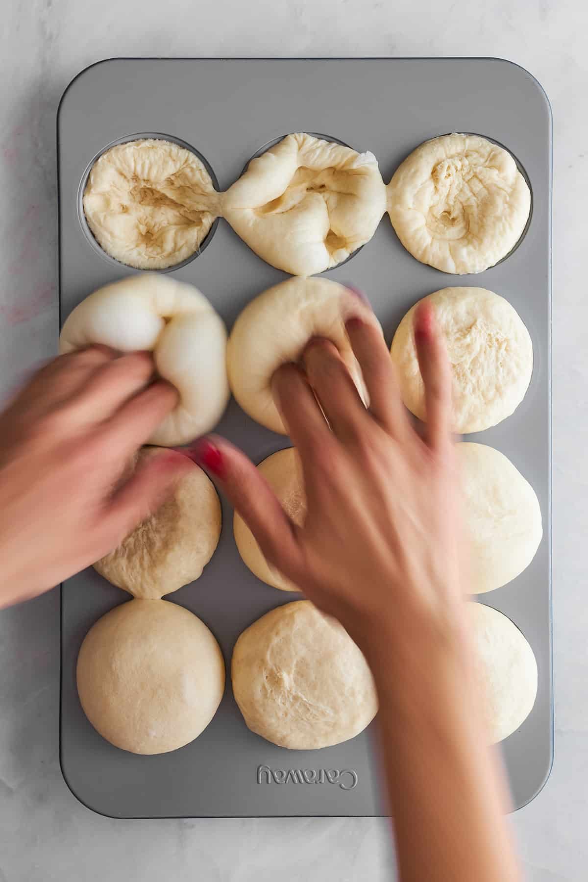 Hands indenting bread rolls in a muffin tin.