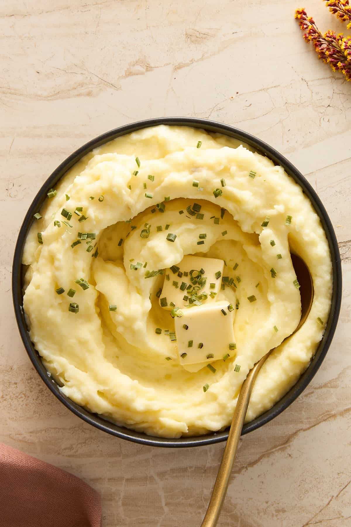 A bowl of creamy mashed potatoes topped with melting butter and chopped chives, with a gold spoon resting inside. The bowl sits on a light-colored marble surface.