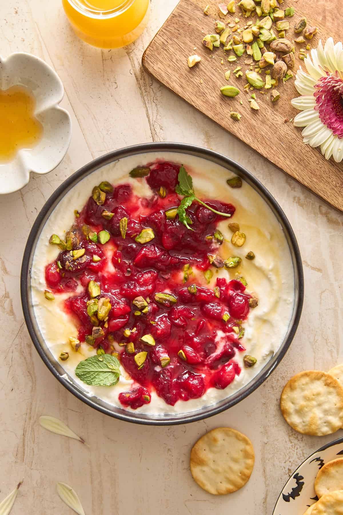 A bowl of silky whipped feta dip topped with red fruit sauce, chopped pistachios, and mint leaves sits on a marble surface. Nearby are crackers, a small dish of honey, orange juice, and a wooden board with chopped pistachios and a pink flower.