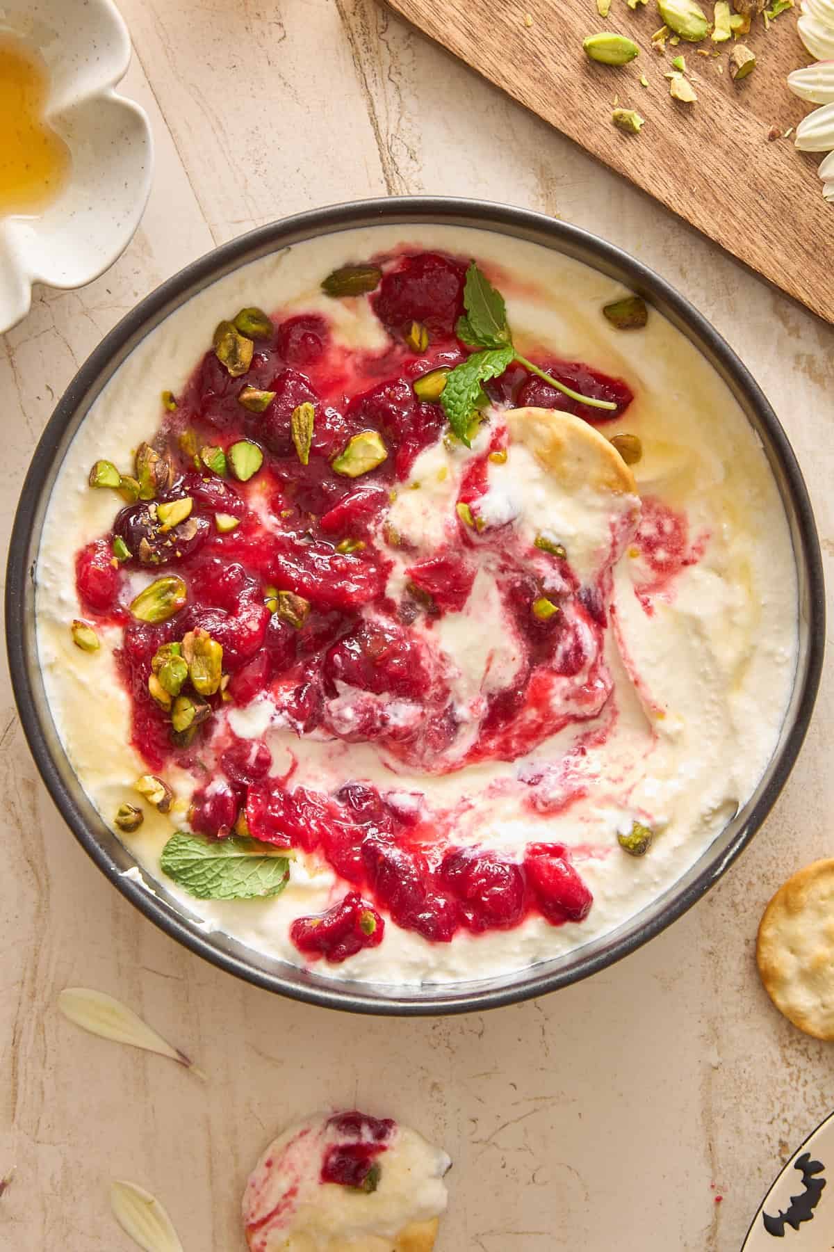A bowl of creamy whipped feta dip topped with a swirl of red fruit compote, chopped pistachios, and fresh mint leaves, set on a light-colored surface with scattered pistachios, mint, and honey nearby.
