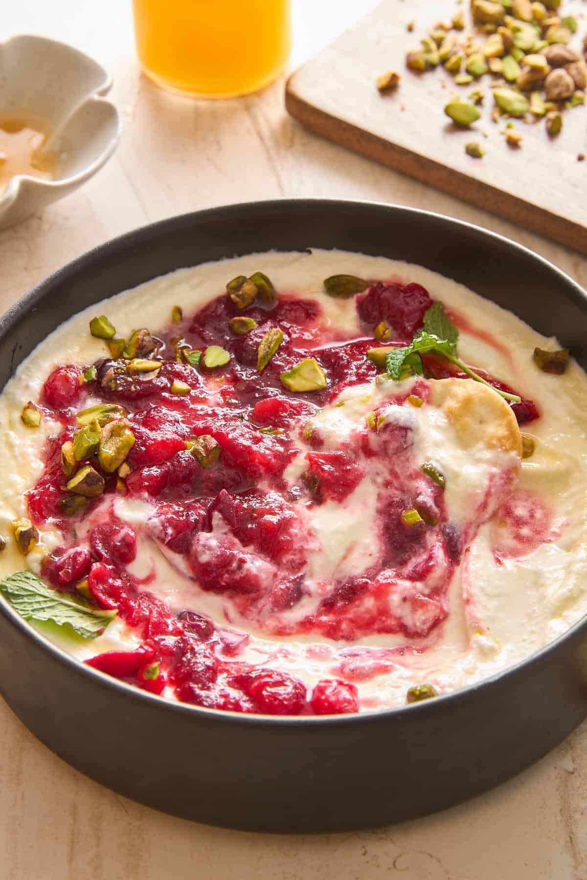 A bowl of creamy yogurt topped with swirling raspberry sauce, chopped pistachios, and fresh mint leaves sits on a light surface, with honey and more pistachios visible in the background.