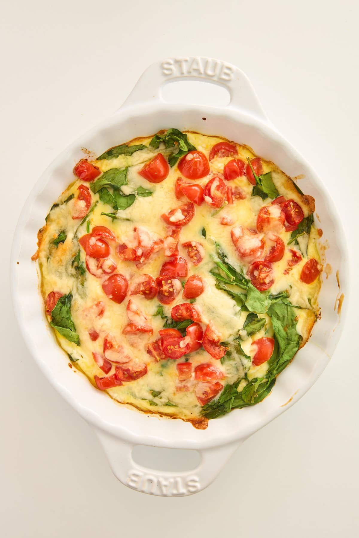 A baked frittata in a white round dish, topped with spinach, chopped cherry tomatoes, and melted cheese, shown from above on a plain light background.