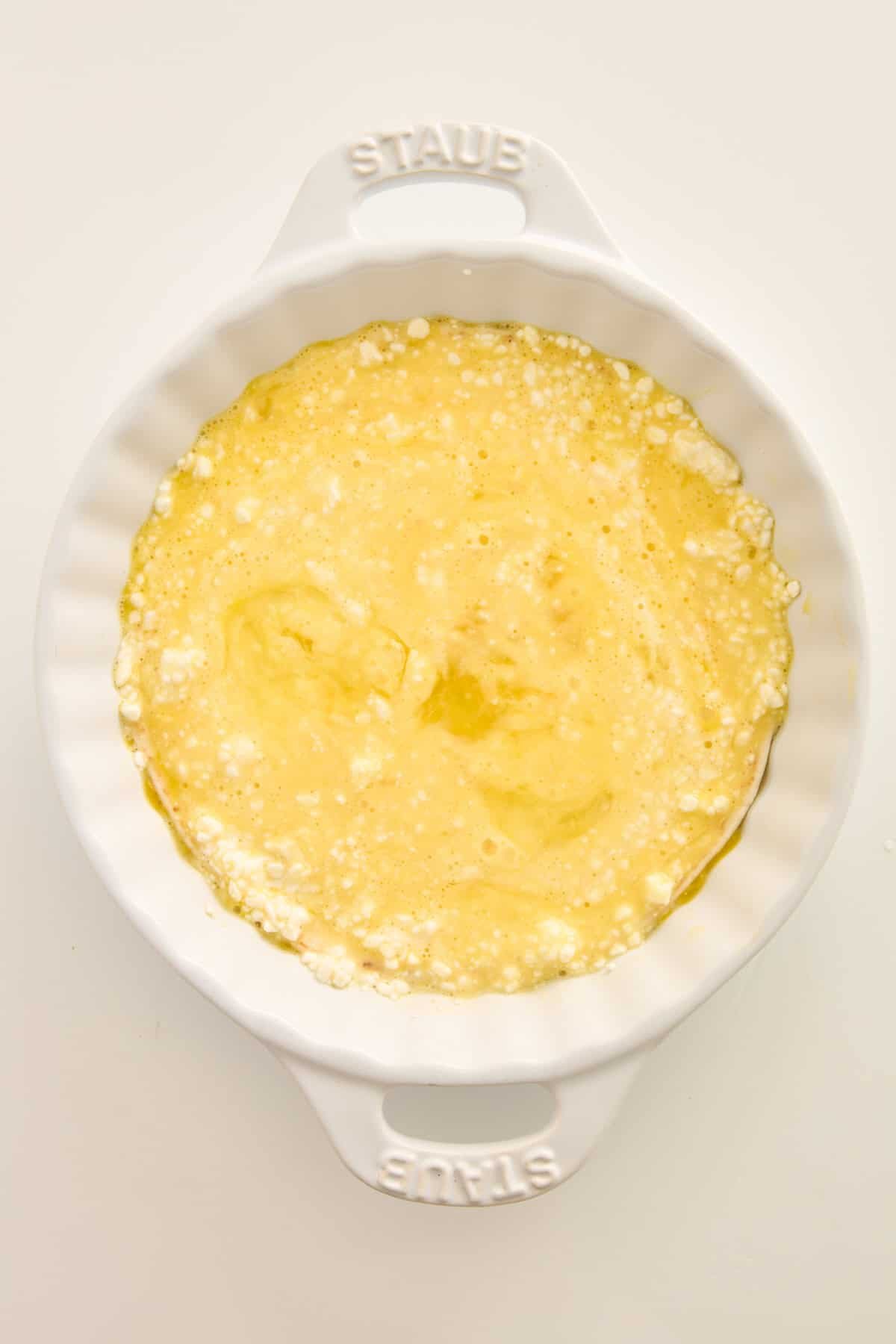 A white ceramic baking dish filled with a yellow, bubbly egg mixture, likely prepared for baking. The dish is placed on a plain white surface.