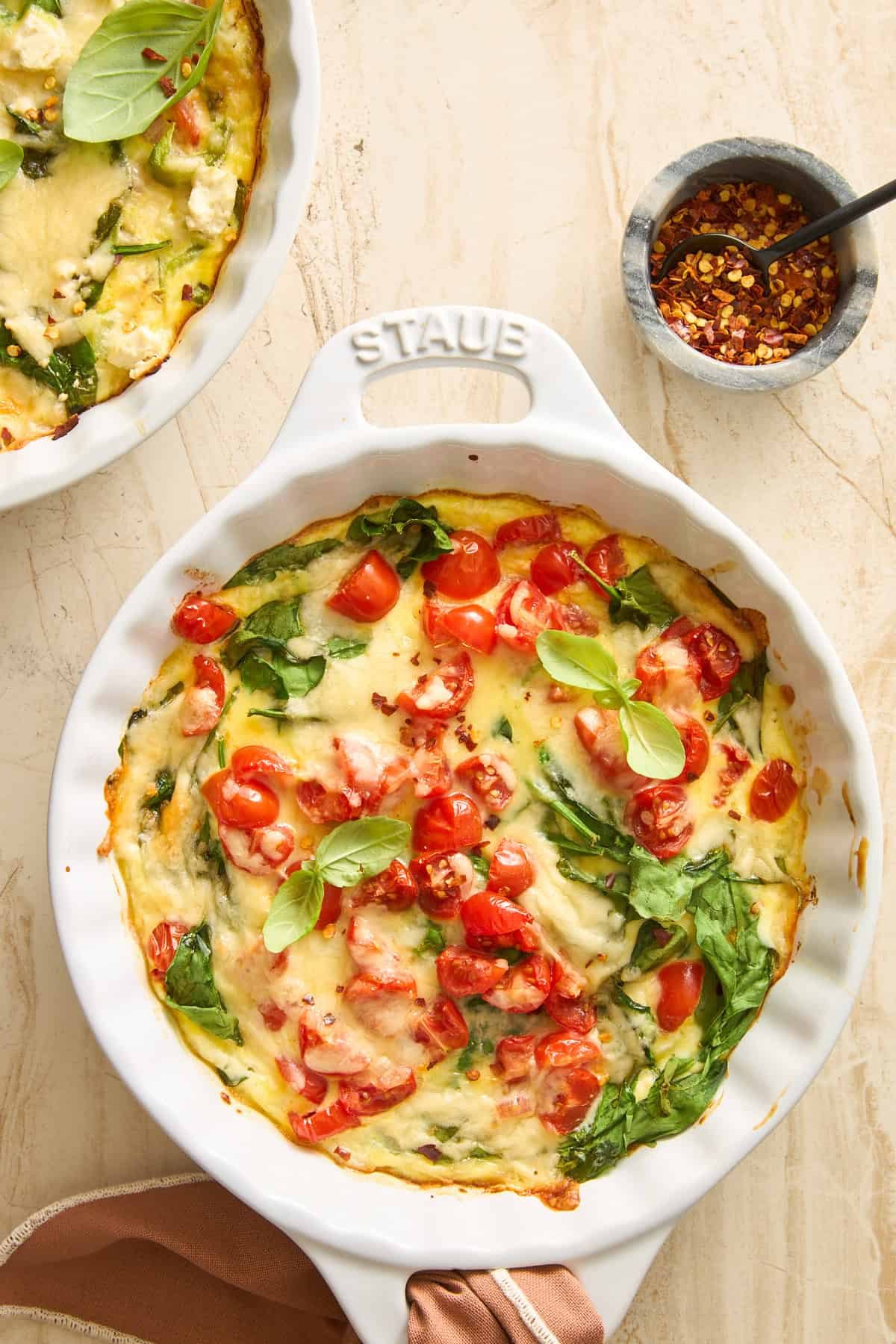 A baked frittata in a round white dish, topped with cherry tomatoes, spinach, melted cheese, and fresh basil leaves. A small bowl of red pepper flakes with a spoon is nearby on a light countertop.