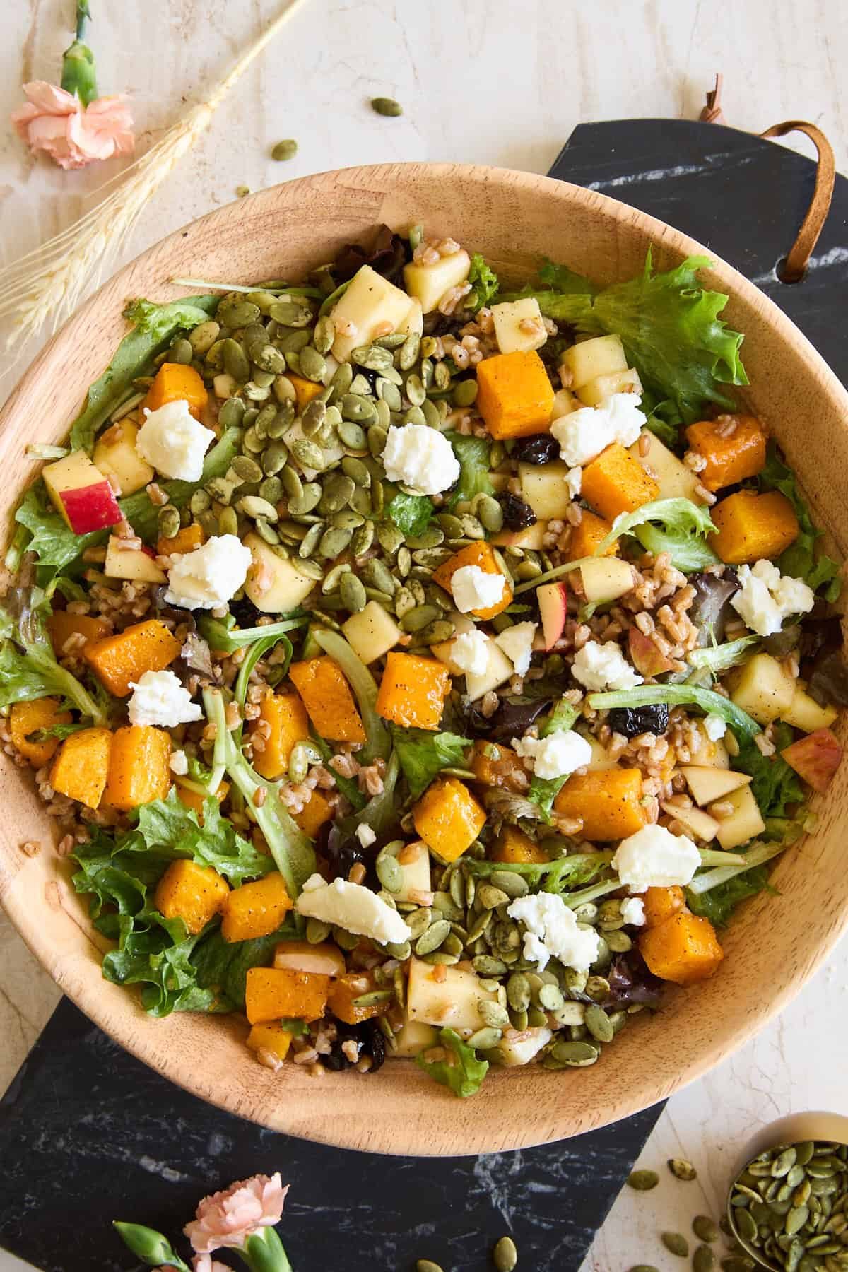 A wooden bowl filled with mixed greens, roasted butternut squash, chopped apples, goat cheese, seeds, and grains, placed on a marble surface with flowers and seeds scattered around.