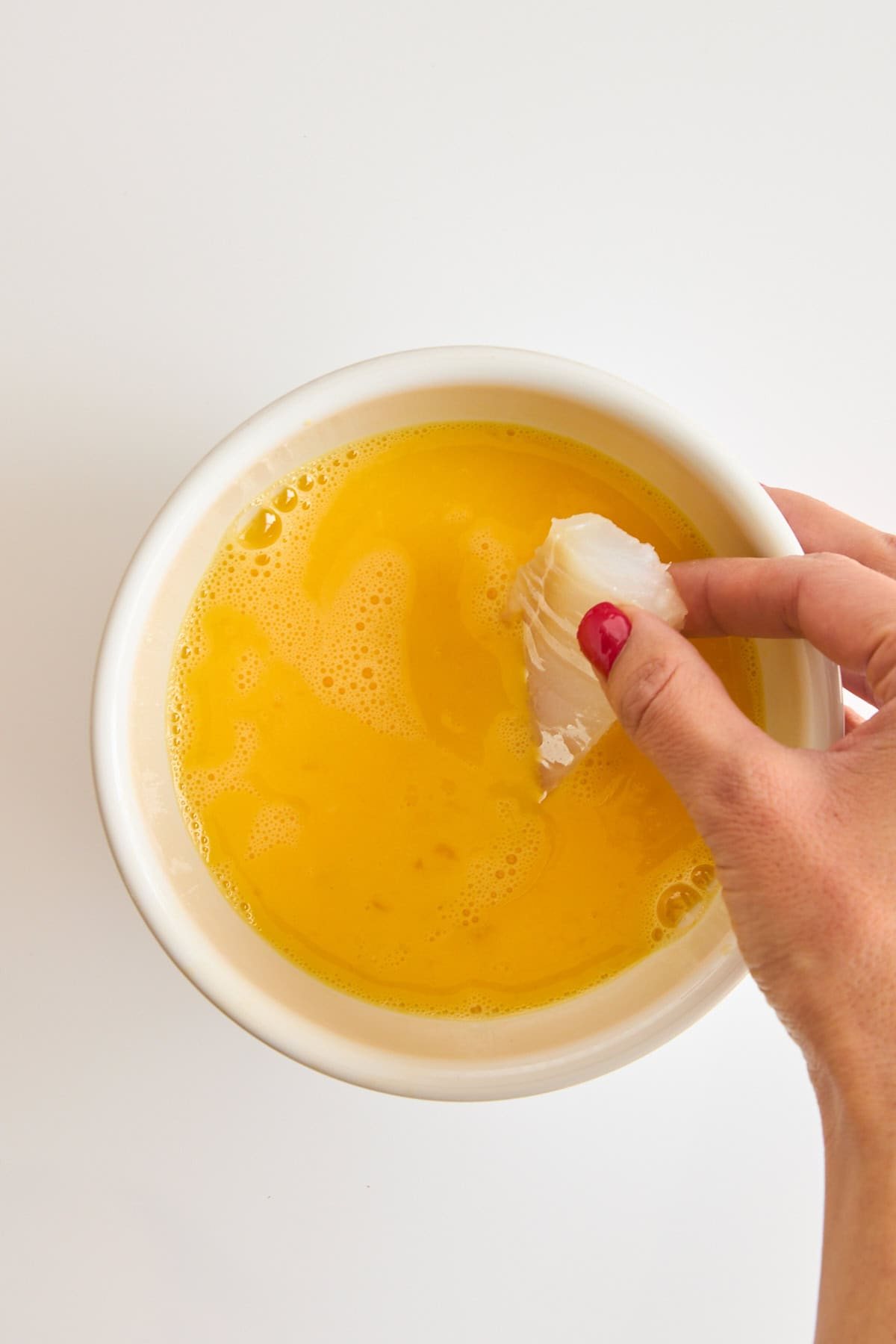 A raw cod piece being dipped into a bowl of whisked eggs.