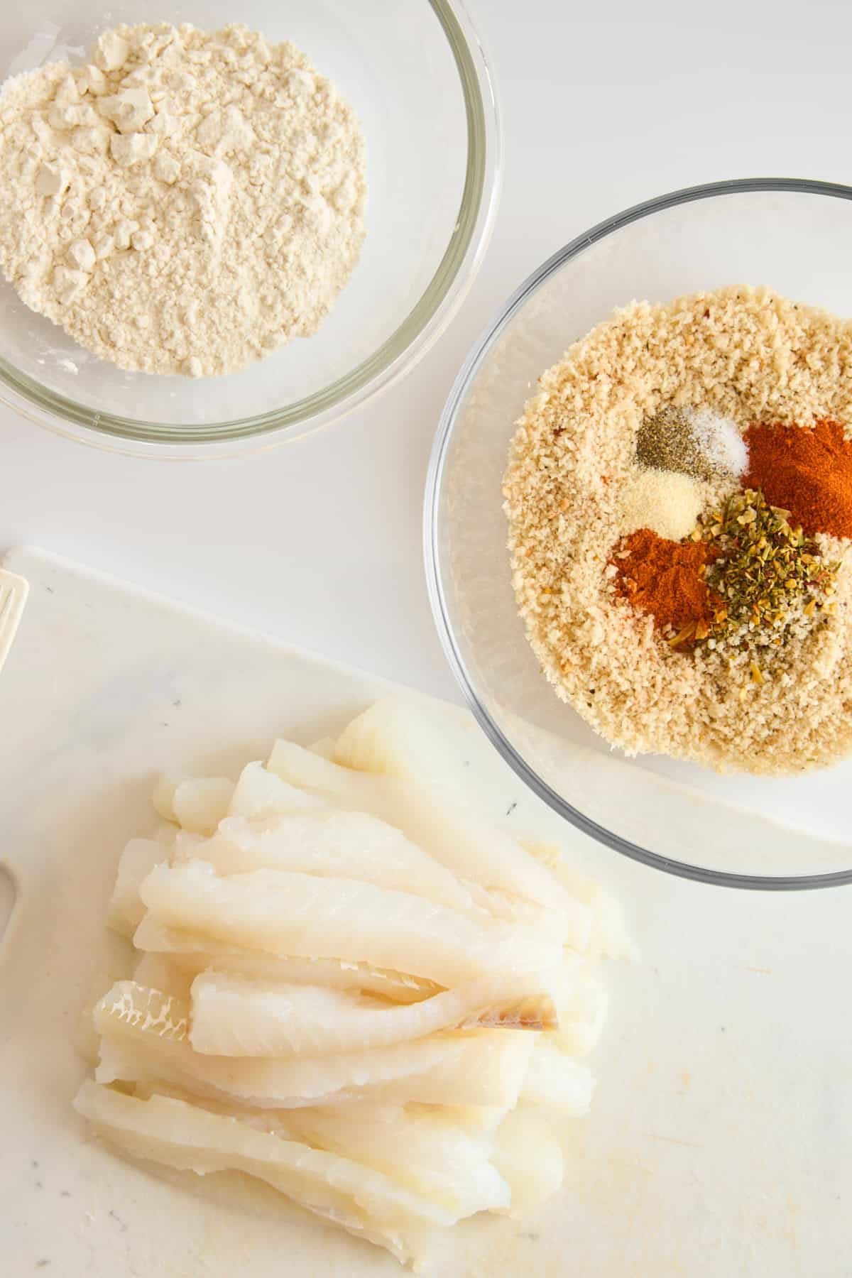 A bowl of flour, a bowl of seasonings, and raw cod filets.