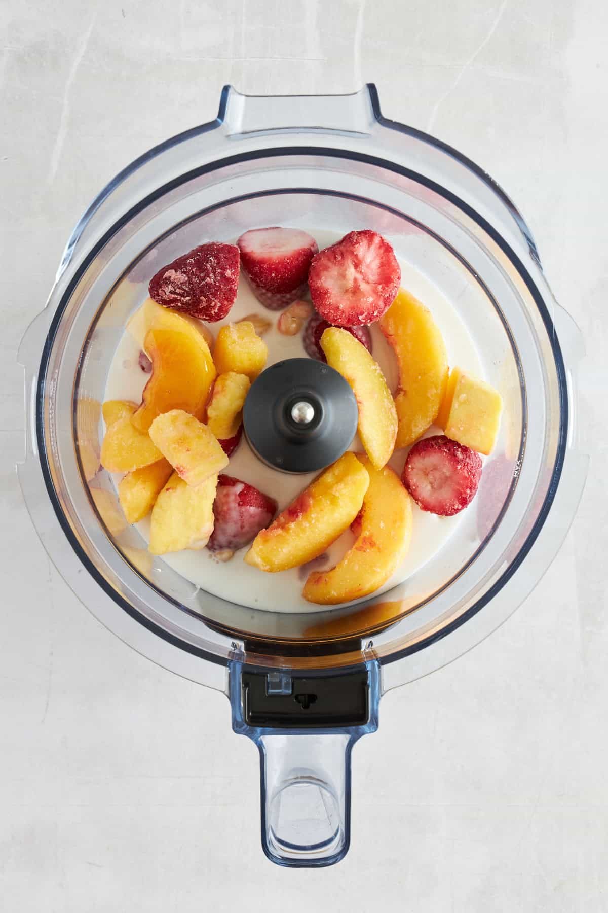 A food processor full of coconut milk, maple syrup, peaches, and strawberries.