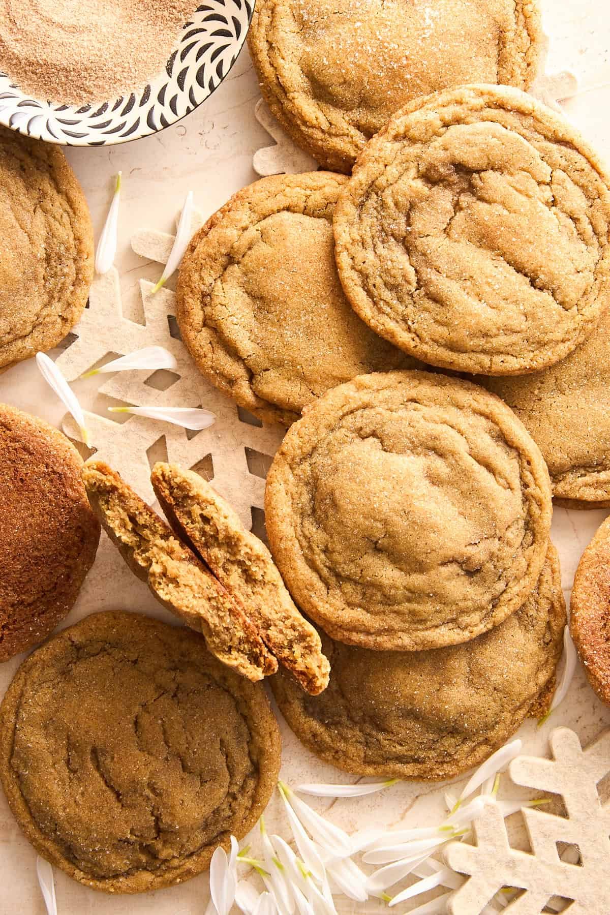 A close-up of several cinnamon sugar cookies on a light surface, with a few flower petals and a decorative paper snowflake. One cookie is broken in half, showing its soft interior. A bowl of cinnamon sugar is in the corner.
