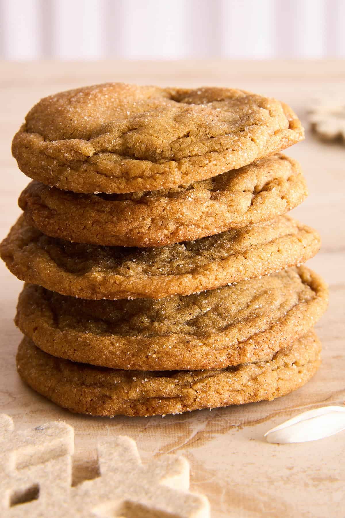 A stack of five golden brown cookies sits on a light wooden surface, with a white snowflake decoration nearby. The cookies appear soft and slightly chewy, with a sugar-coated texture.