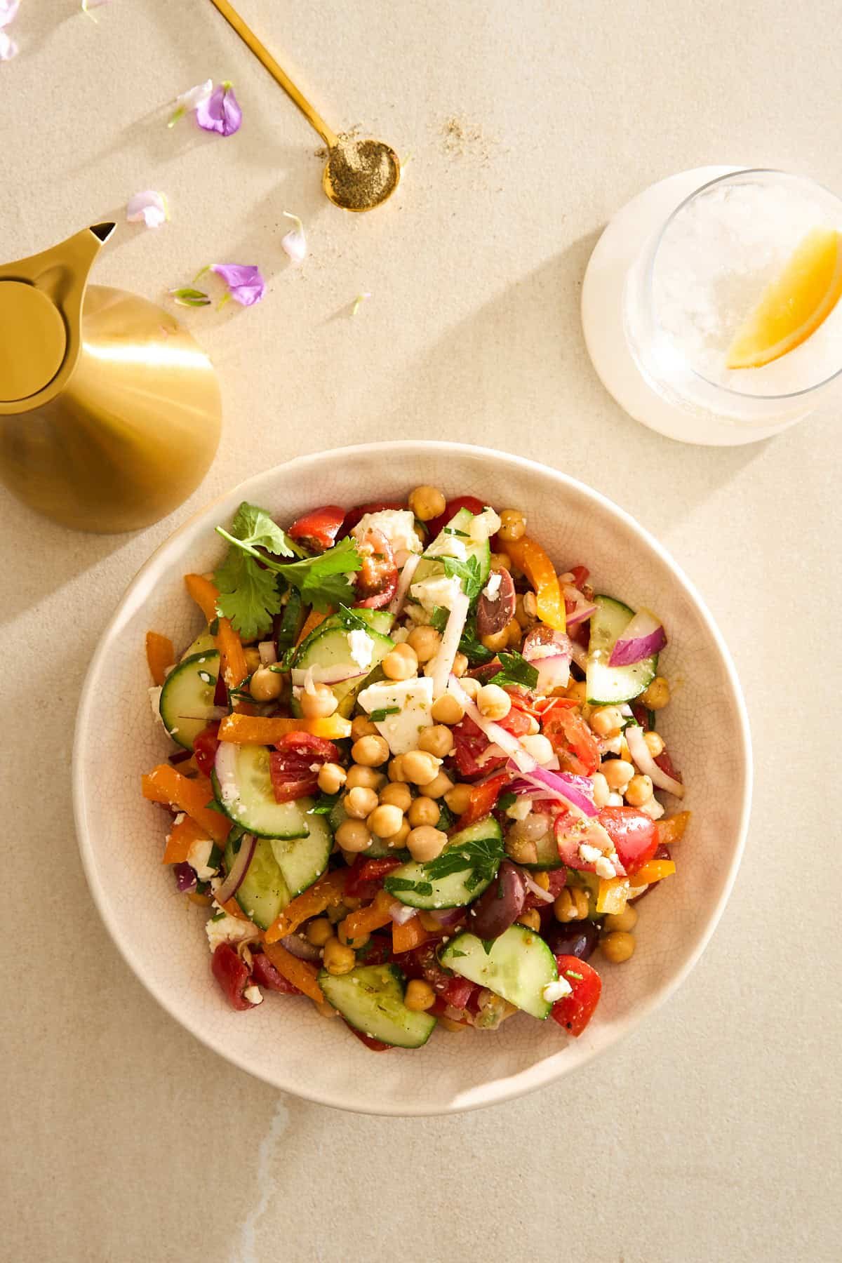 A bowl of colorful chickpea salad with cucumbers, tomatoes, olives, red onion, and herbs on a light surface, next to a gold pitcher, a spoon with spices, and a glass with ice and a lemon twist.