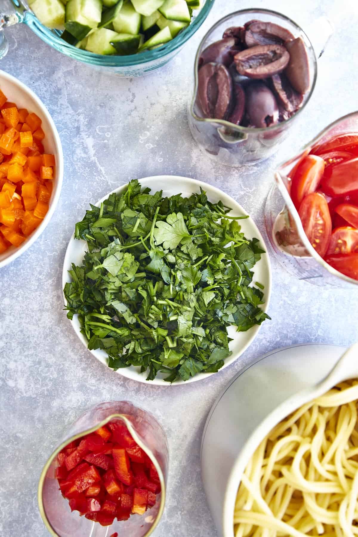Bowls of kalamata olives, halved tomatoes, chopped red and orange bell peppers, cooked spaghetti noodles, and fresh parsley.