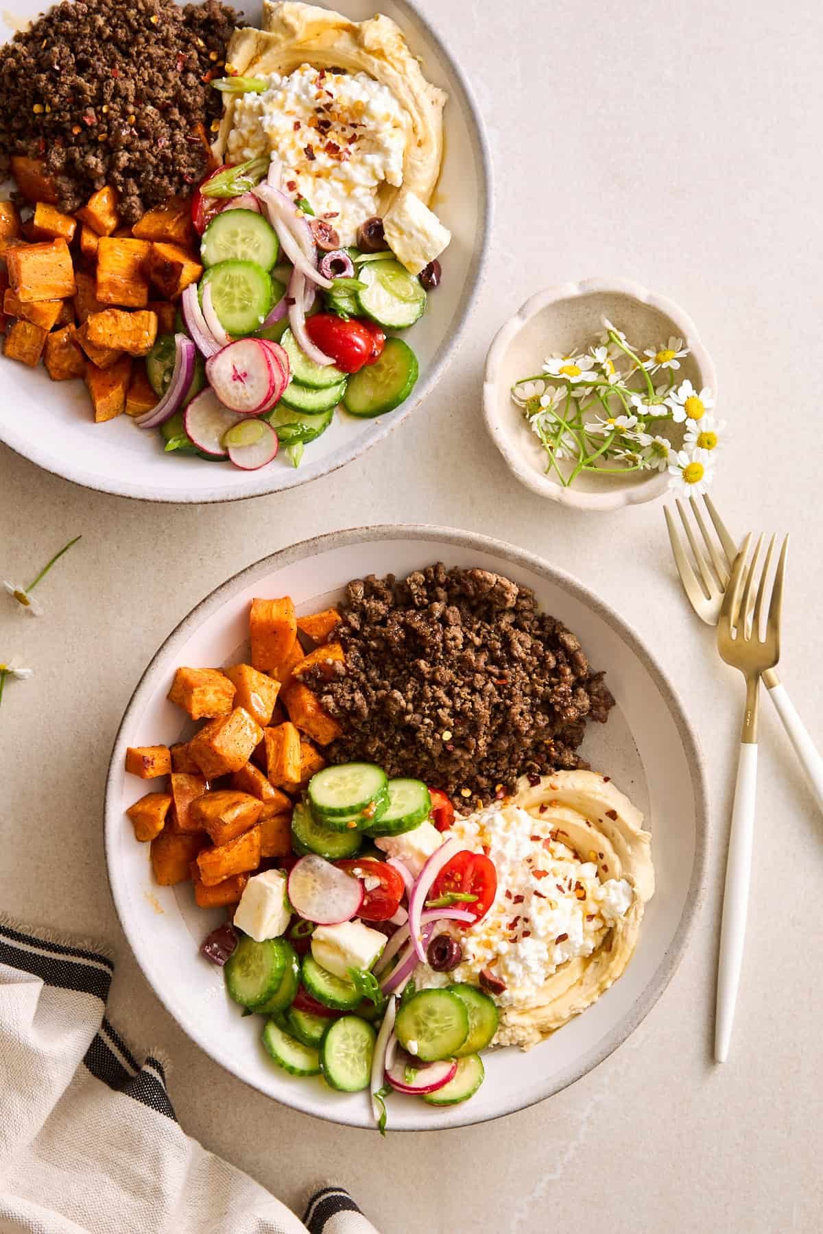 Two ground beef bowls with beef, roasted sweet potatoes, fresh veggies, feta, hummus, and cottage cheese.