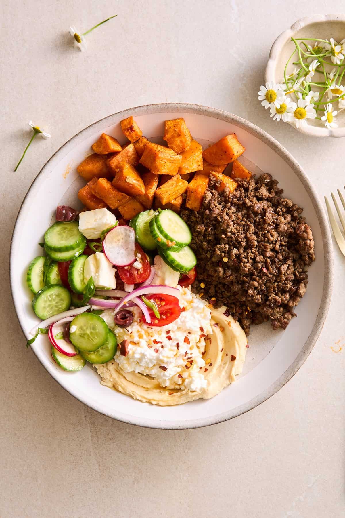 A ground beef bowl with beef, roasted sweet potatoes, fresh veggies, feta, hummus, and cottage cheese.