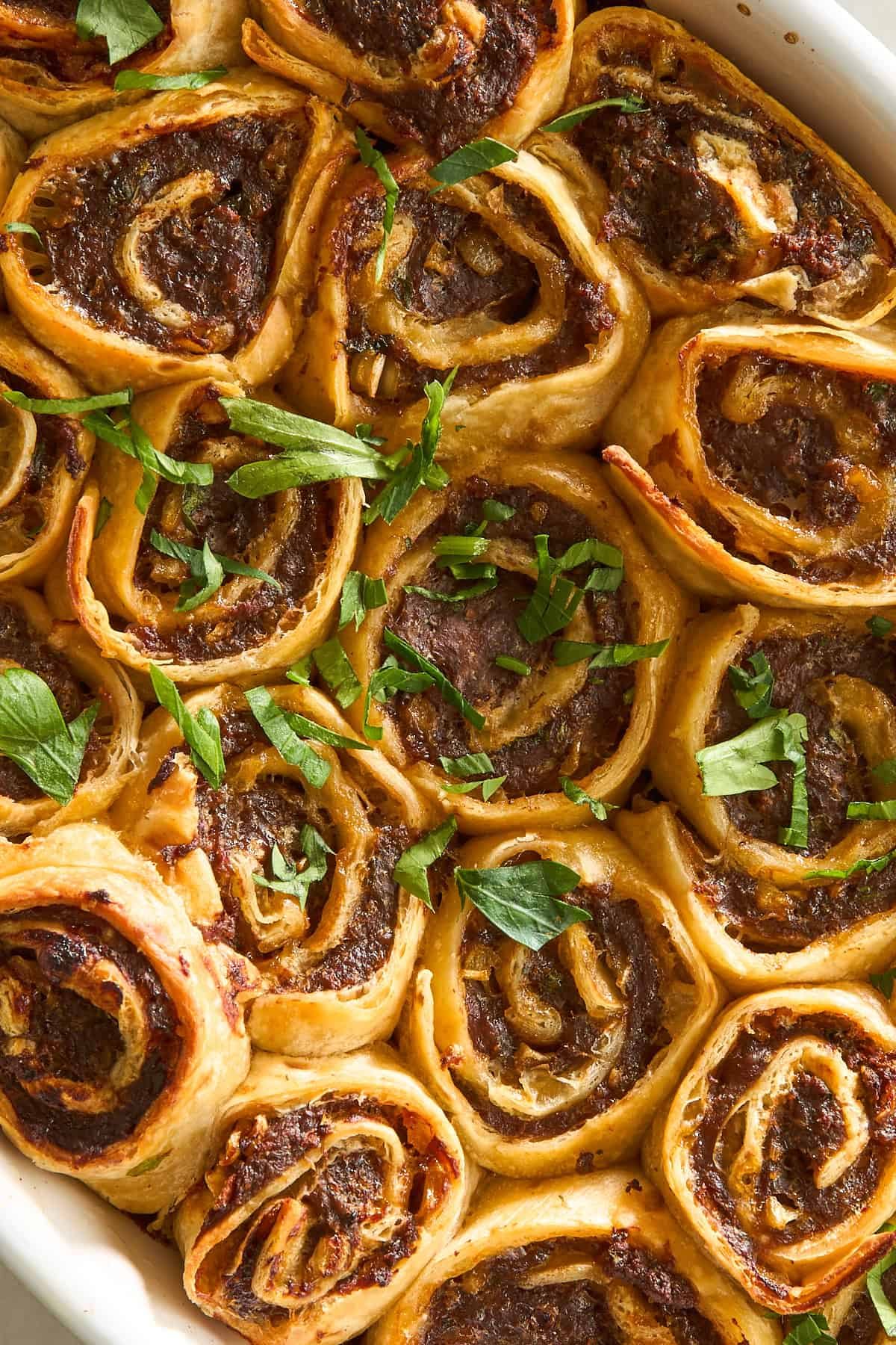Golden-brown pinwheel pastries filled with a savory meat mixture, baked in a dish and garnished with fresh chopped parsley. The rolls are closely packed and have a crispy, flaky texture.