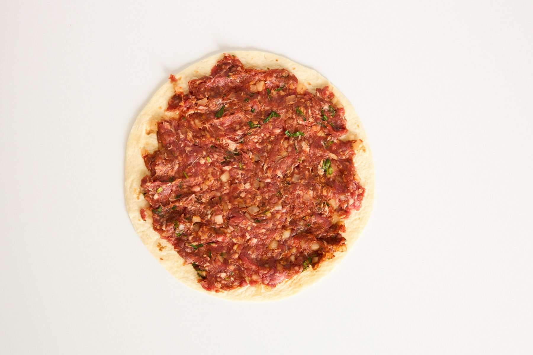 A round piece of flatbread topped with a raw, finely minced meat mixture, spread evenly to the edges, on a plain white background.