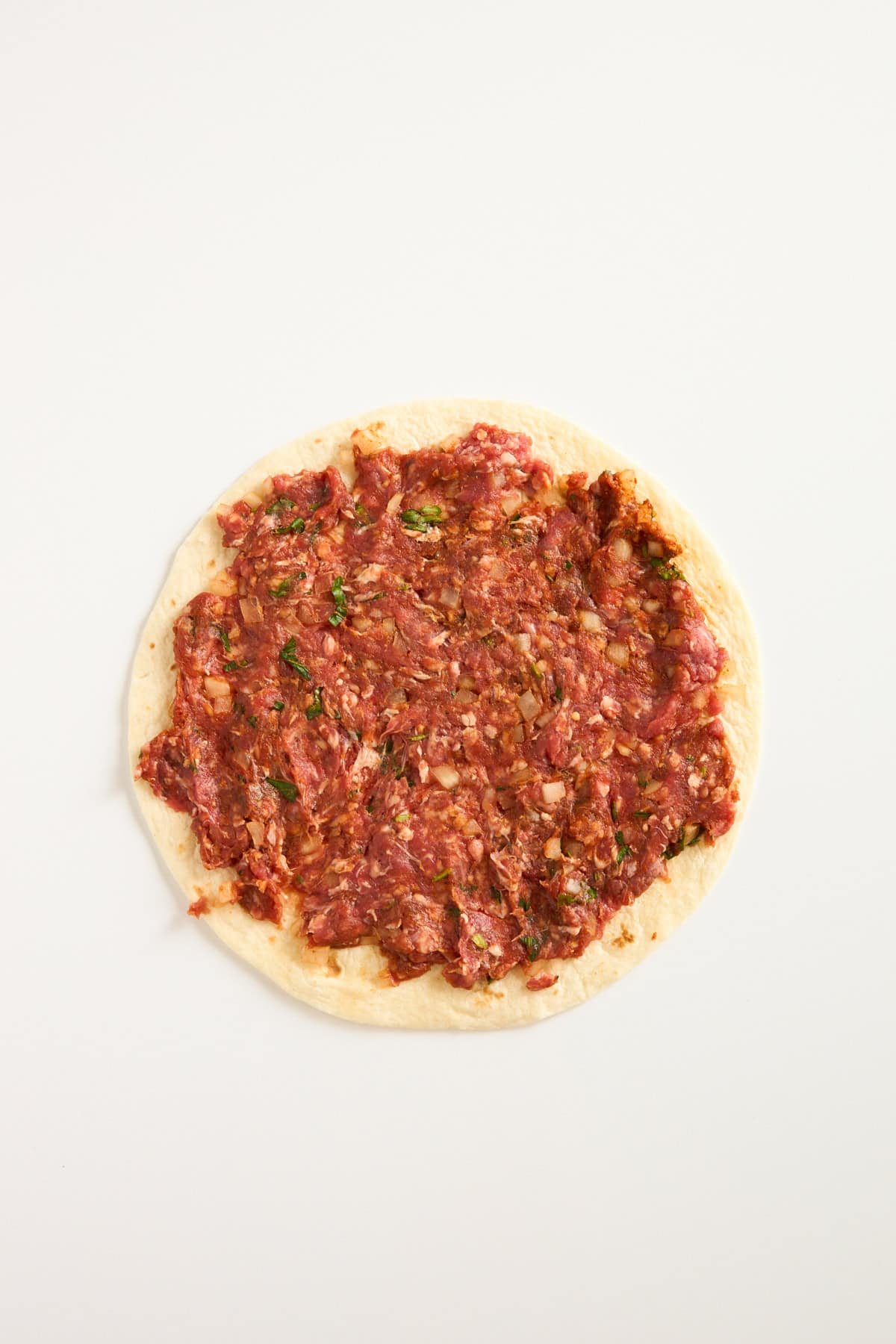 A round piece of flatbread topped with a raw, finely minced meat mixture, spread evenly to the edges, on a plain white background.