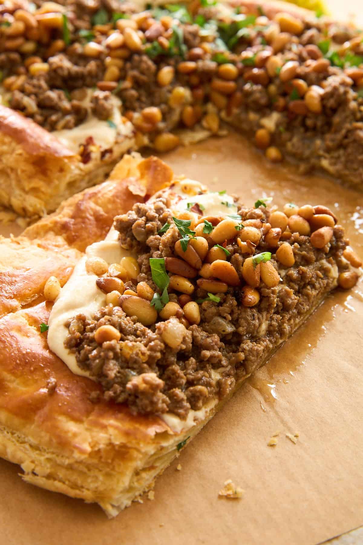 A close-up of a rectangular pastry topped with seasoned ground meat, toasted pine nuts, and chopped parsley, resting on parchment paper. One piece is cut and slightly pulled away from the rest.