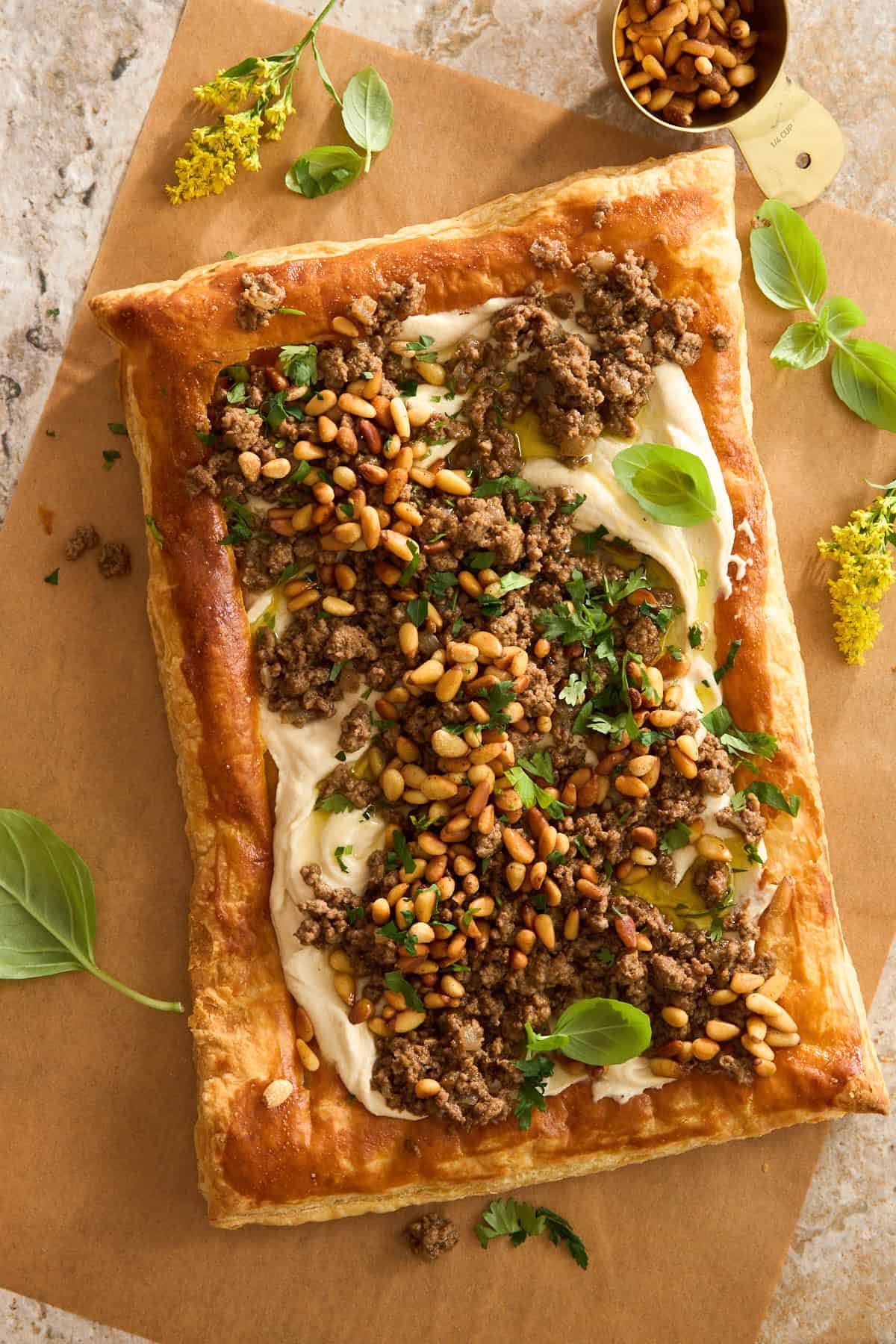 A rectangular puff pastry tart topped with silky spread, cooked ground meat, toasted pine nuts, fresh herbs, and basil leaves, placed on parchment paper with a gold measuring cup and yellow flowers nearby.