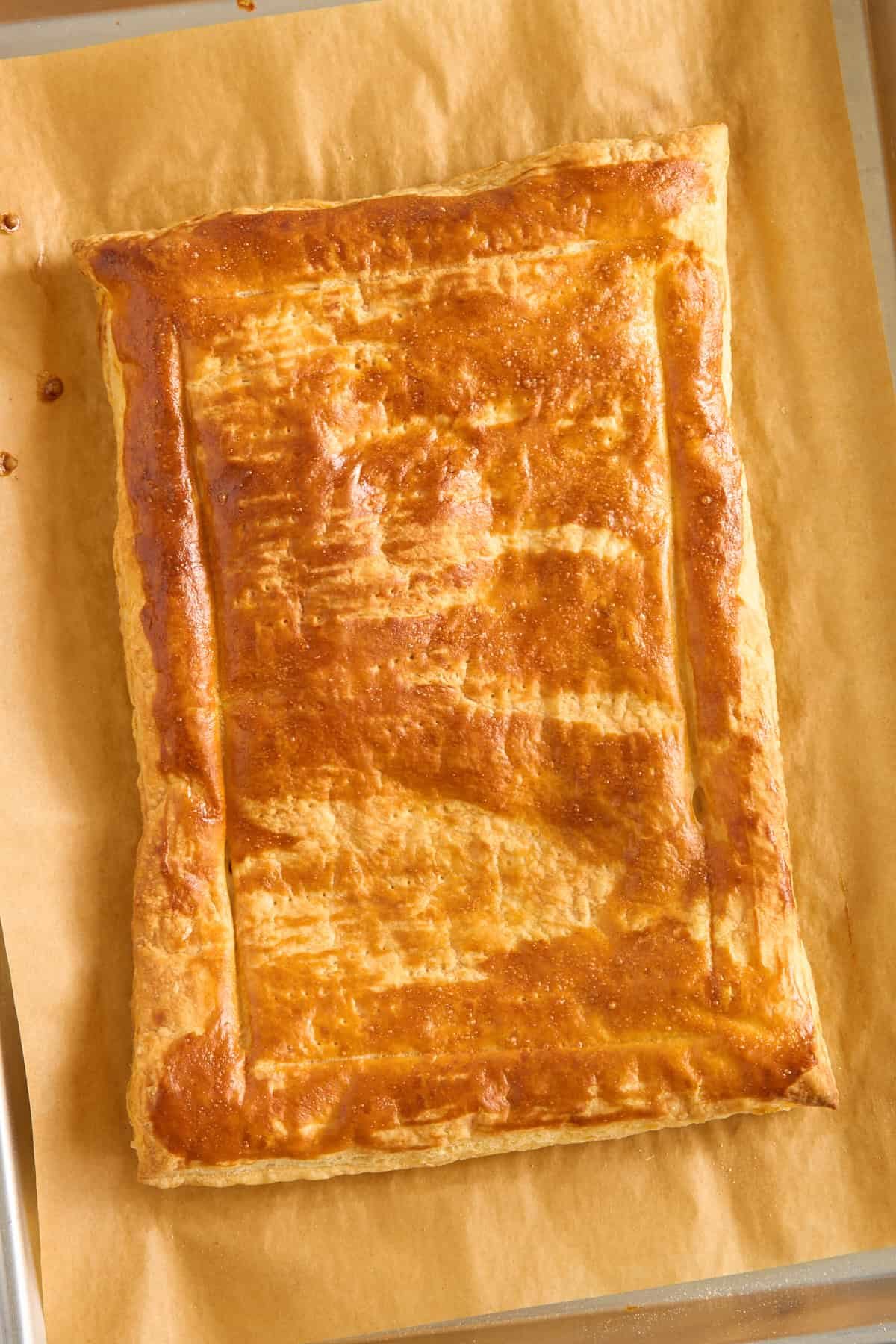 A rectangular golden-brown pastry with a shiny, flaky crust sits on a sheet of parchment paper. The edges are crimped, and the top shows light bake marks.