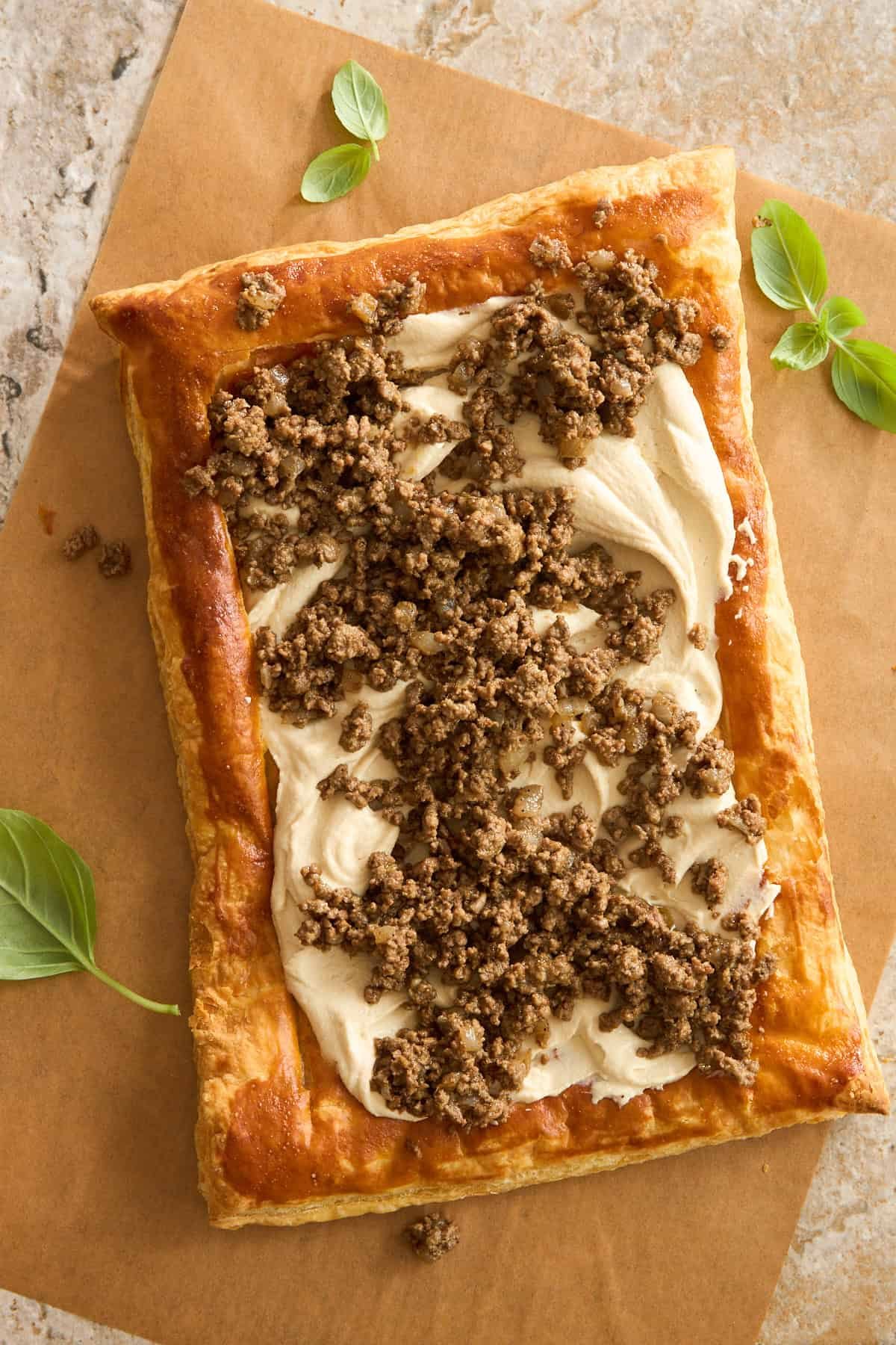 A rectangular puff pastry topped with a creamy spread and crumbled cooked sausage sits on brown parchment paper, garnished with fresh basil leaves on the side.