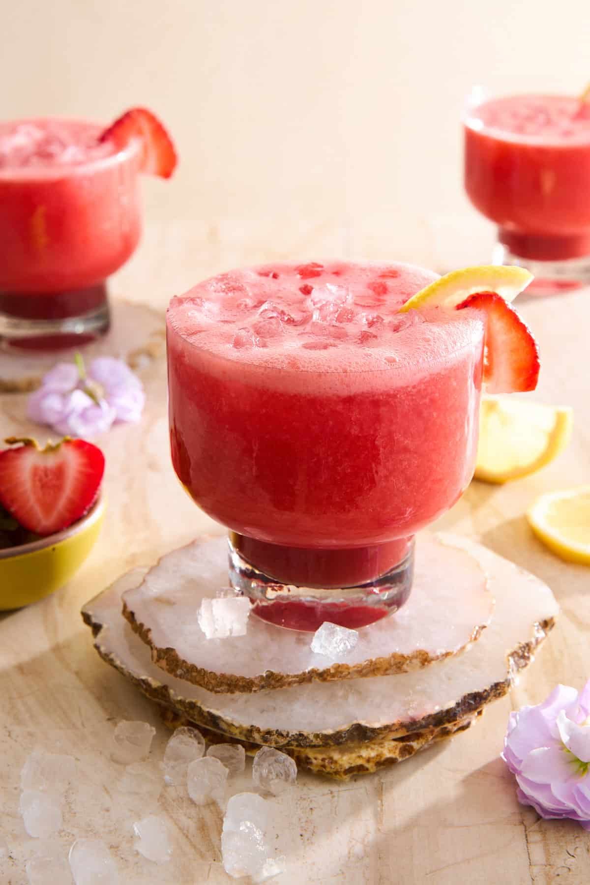 A glass of pink strawberry lemonade with ice, garnished with a lemon slice and strawberry, sits on stone coasters.