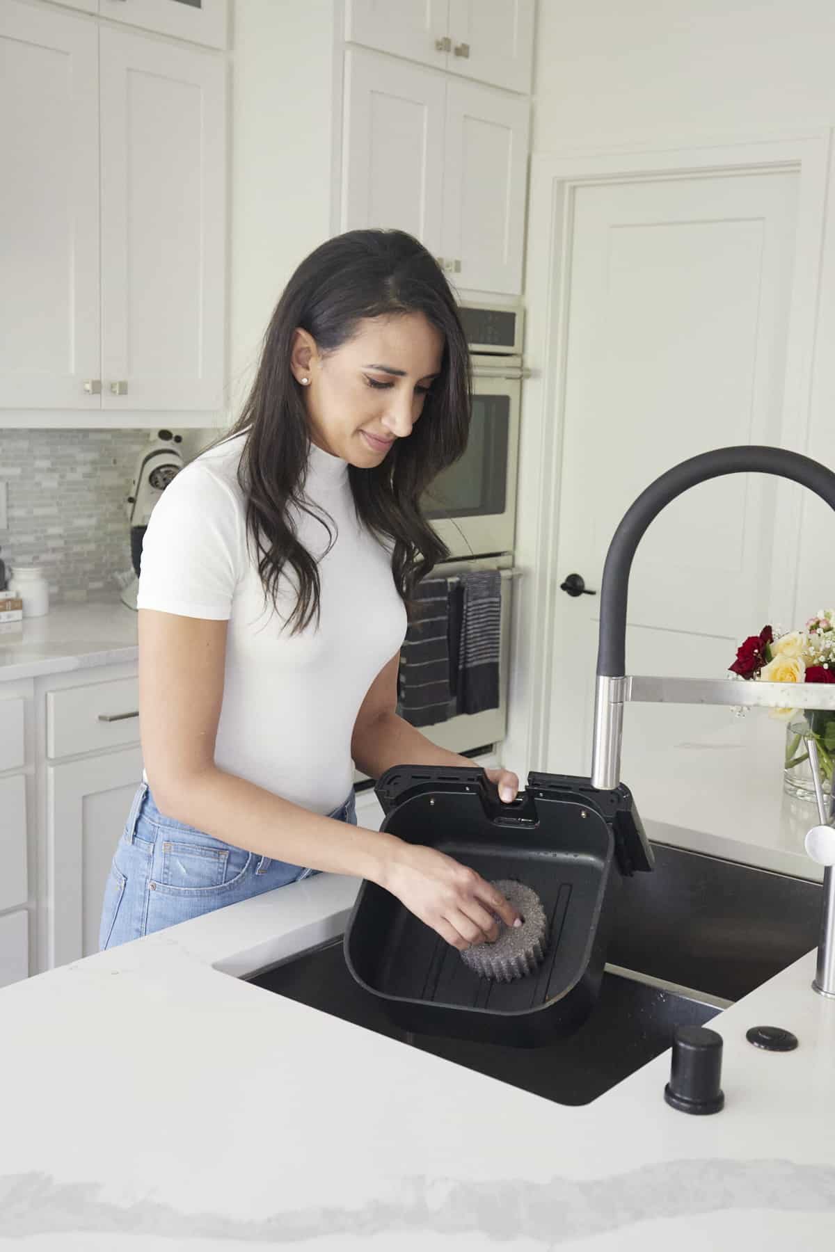 A woman using a Scrub Daddy brush to clean the basket of an air fryer ina. sink.