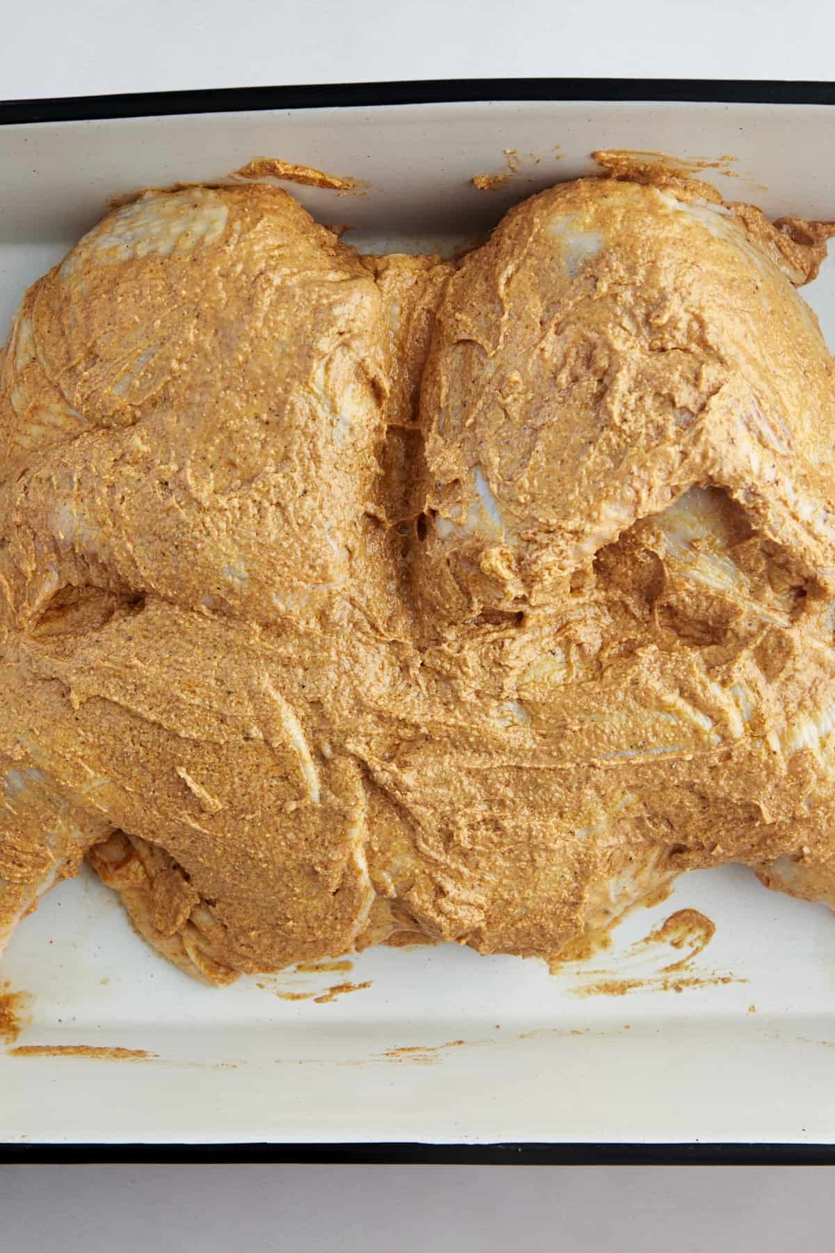 A raw butterflied chicken covered with a spiced yogurt marinade in a baking dish.