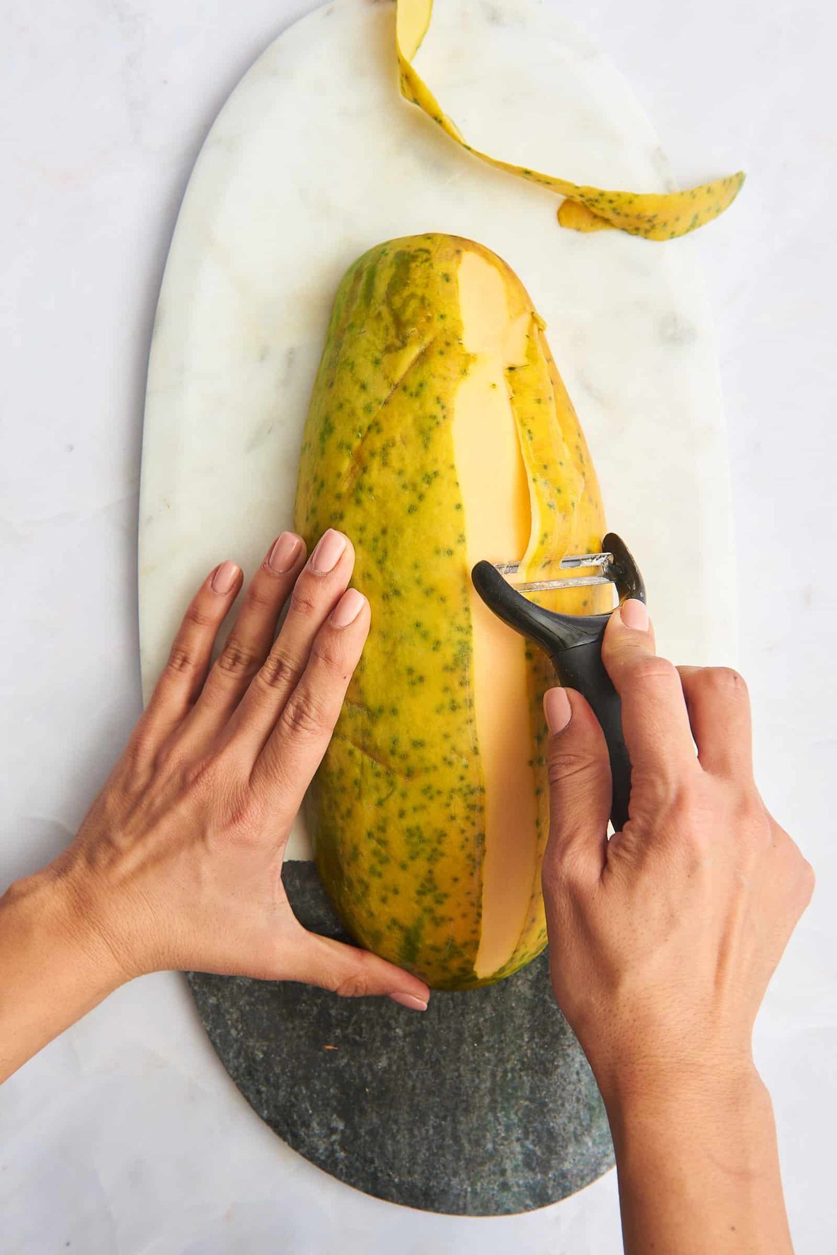 A hand using a vegetable peeler to remove the skin from a papaya.