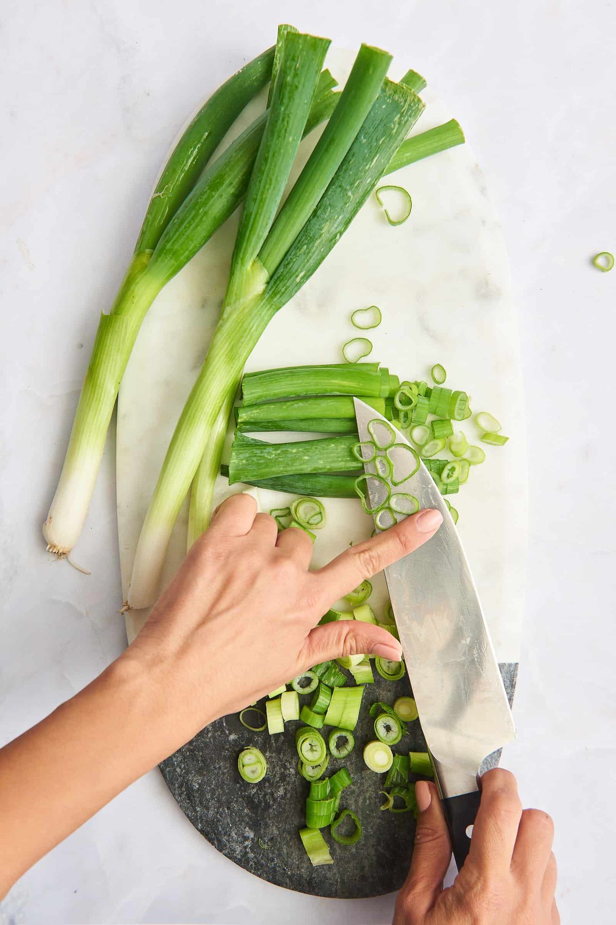 A knife with chopped scallions over a cutting board with whole and chopped green onions.