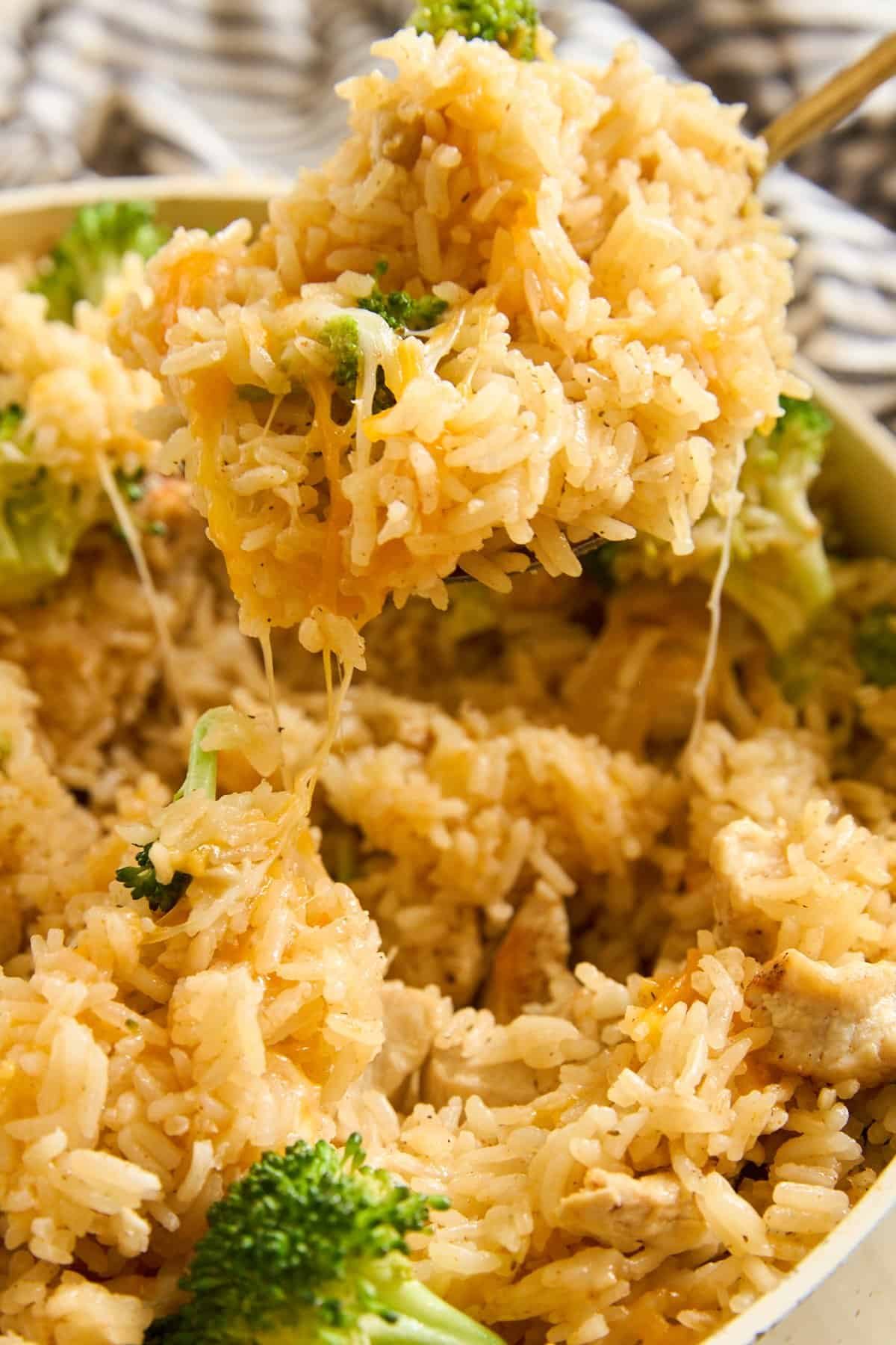 Close-up of a spoon lifting cheesy rice with broccoli out of a dish, with melted cheese stretching from the spoonful. Rice and broccoli are mixed together, and cheese is visible throughout the dish.