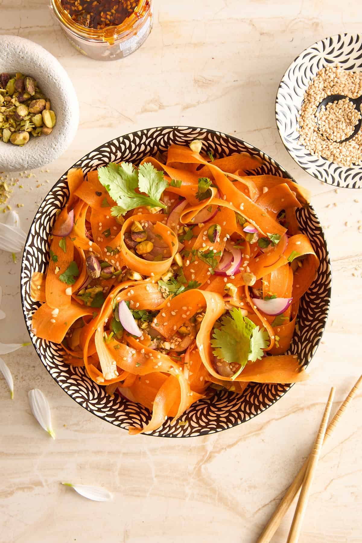 A bowl of carrot ribbon salad garnished with cilantro, red onion, sesame seeds, and pistachios sits on a light surface surrounded by a dish of sesame, a jar of chili flakes, and chopsticks.