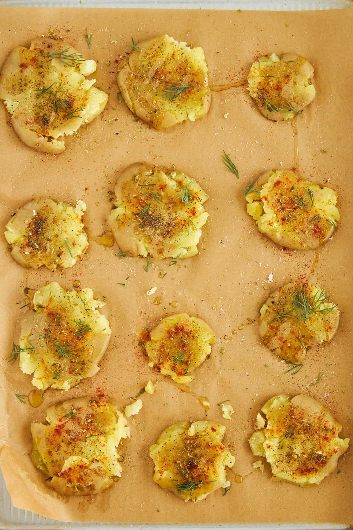 Uncooked smashed potatoes topped with oil and seasonings on a baking sheet.
