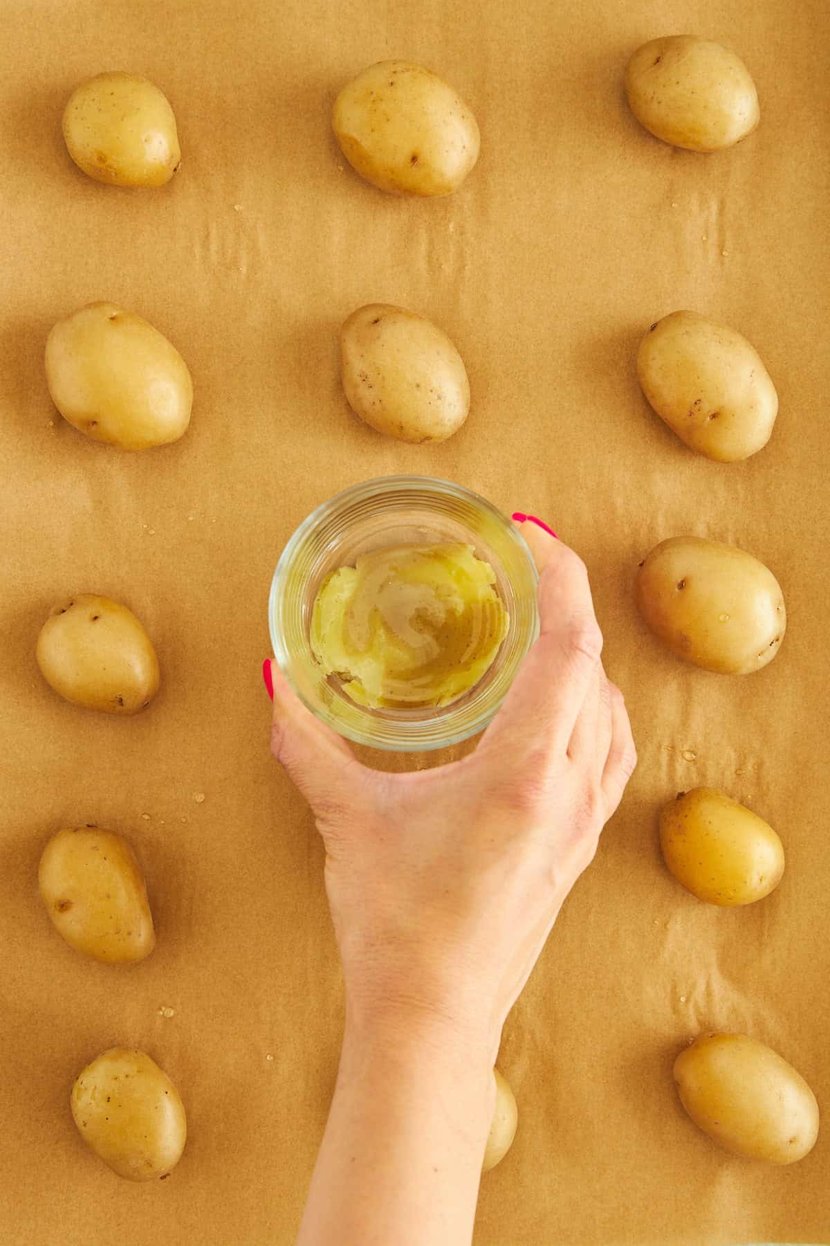 A hand using a cup to smash potatoes on a baking sheet.