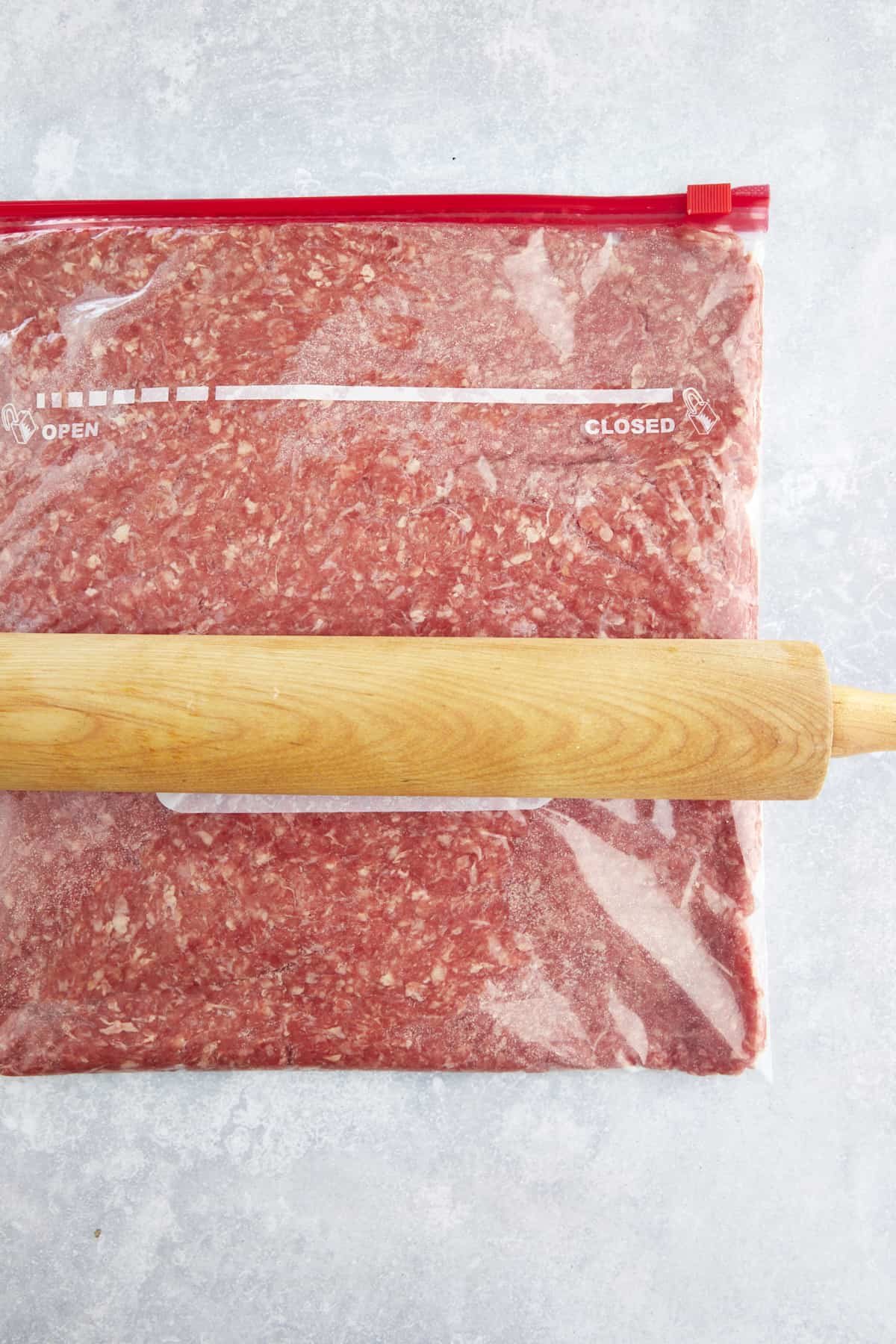 A rolling pin flattening raw ground beef in a sealable bag.