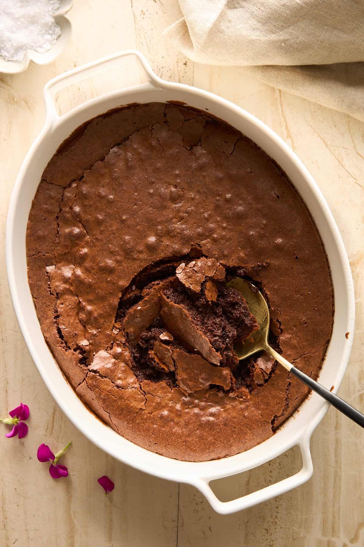 Oval white baking dish with a cracked, rich, baked chocolate dessert. A spoon is scooping out a portion from the center. Purple flower petals and a bowl of white powder are beside the dish on a light surface.