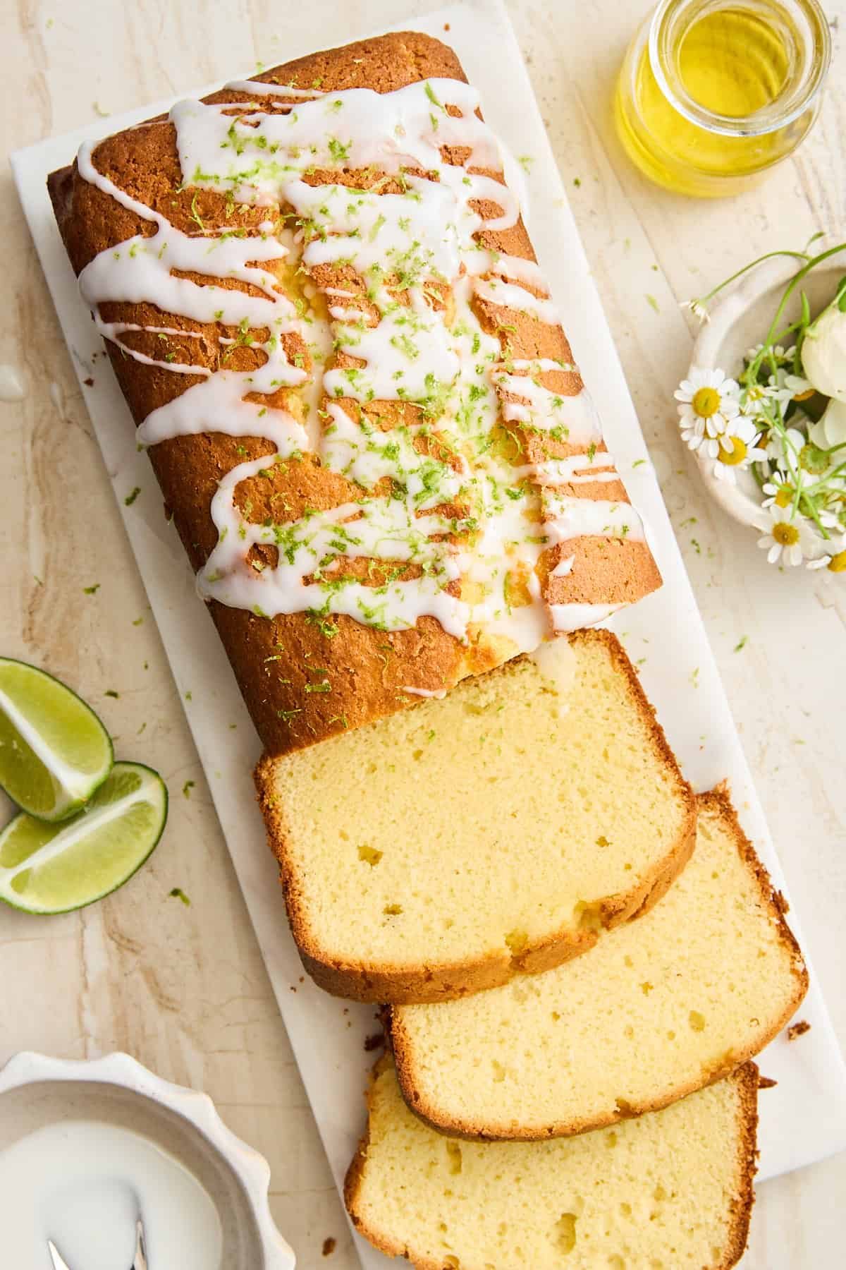 A loaf of glazed pound cake, drizzled with white icing and topped with lime zest, sits on a marble board. Several slices are cut, with lime wedges and a small jar of oil nearby, and flowers as decoration.
